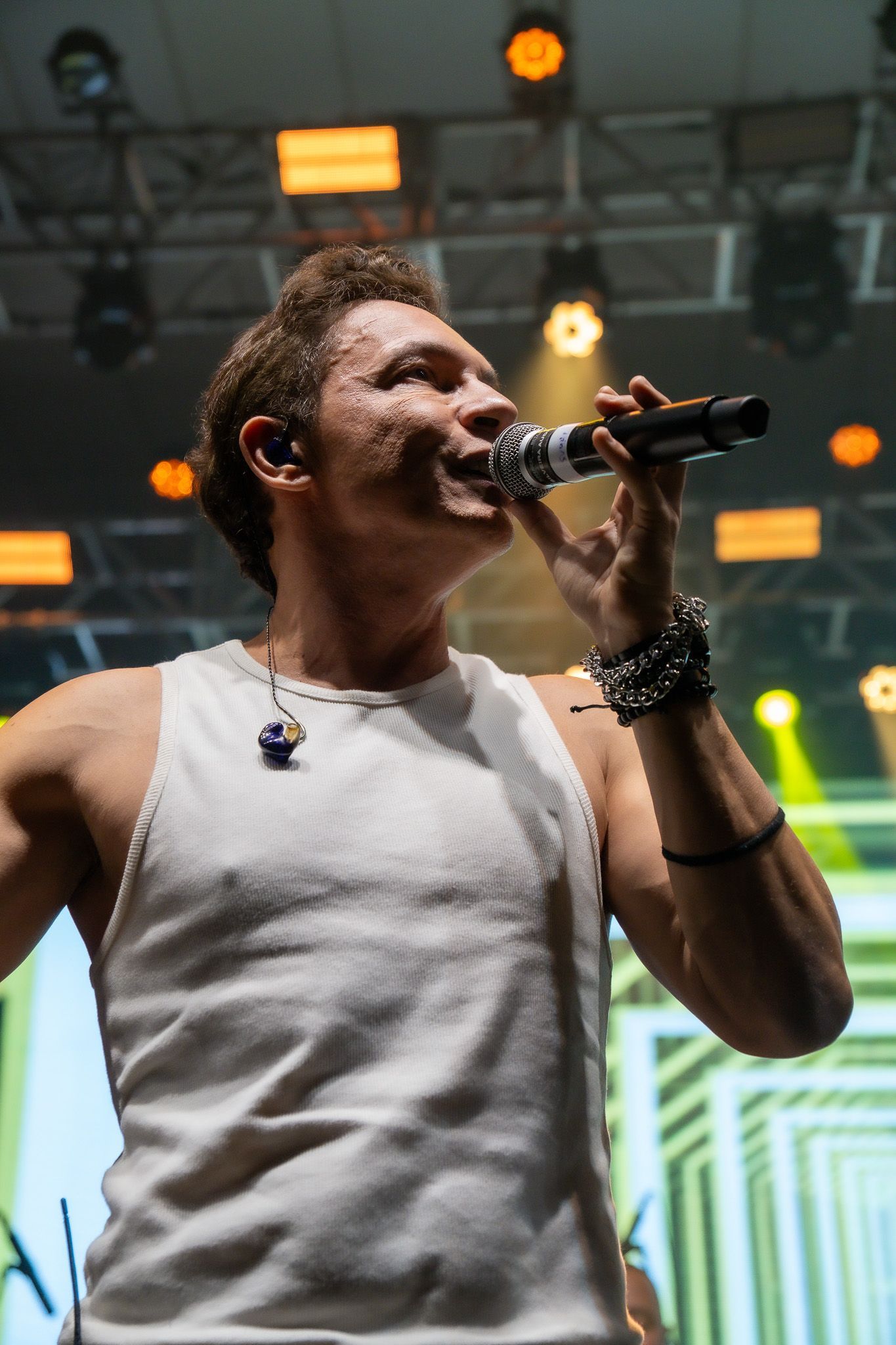 Um artista vestindo uma regata branca canta em um microfone em um palco com iluminação forte ao fundo.