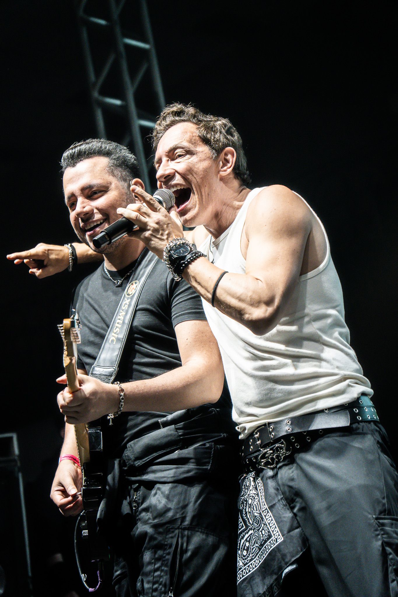 Dois artistas se apresentam em um palco escuro; um toca guitarra e sorri enquanto o outro canta com entusiasmo em um microfone.