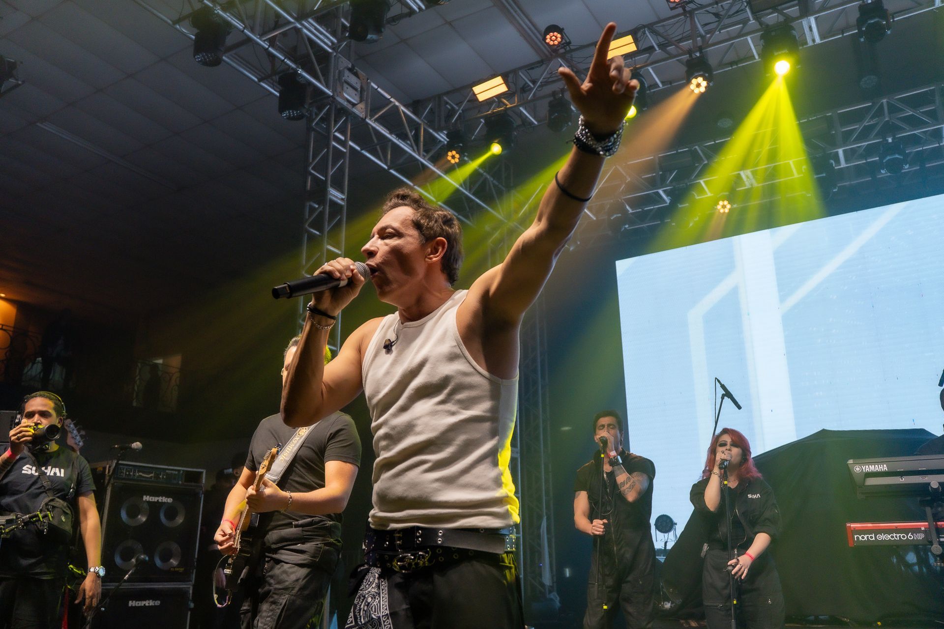 Um artista vestindo uma regata branca canta em um microfone no palco, com o braço levantado, acompanhado por uma banda