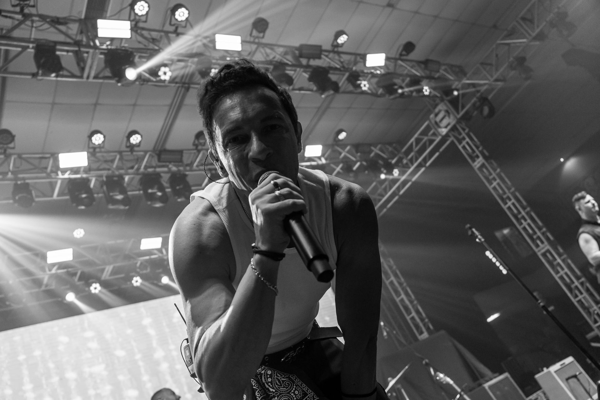 Nesta foto em preto e branco, um músico canta ao microfone em um palco de concertos iluminado por holofotes.