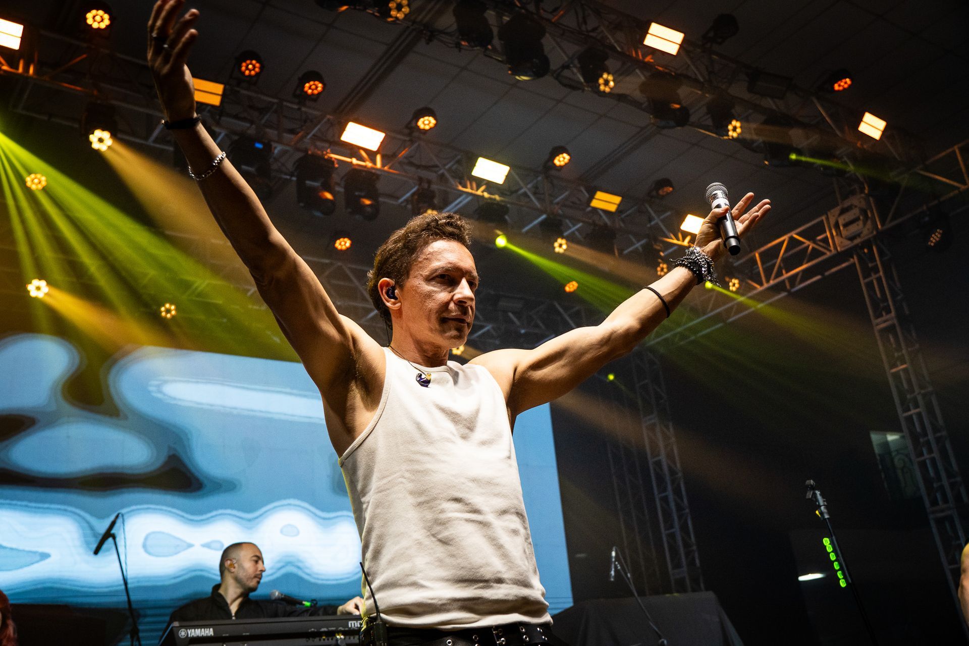 Um artista vestindo uma regata branca está no palco com os braços erguidos, segurando um microfone em meio às luzes do palco.