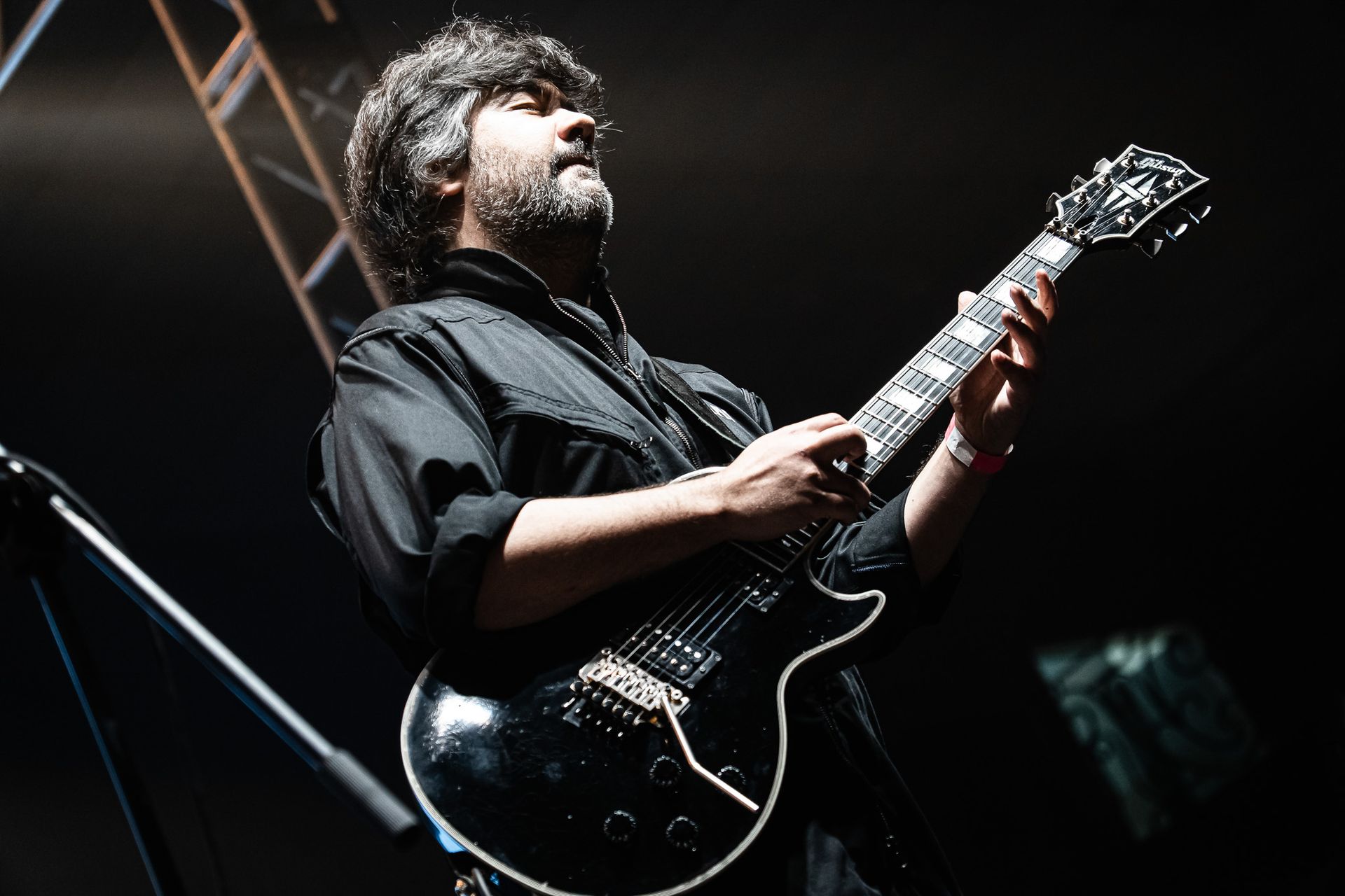 Um artista de cabelos cacheados se apresentando no palco, tocando uma guitarra elétrica preta enquanto olha para a luz.