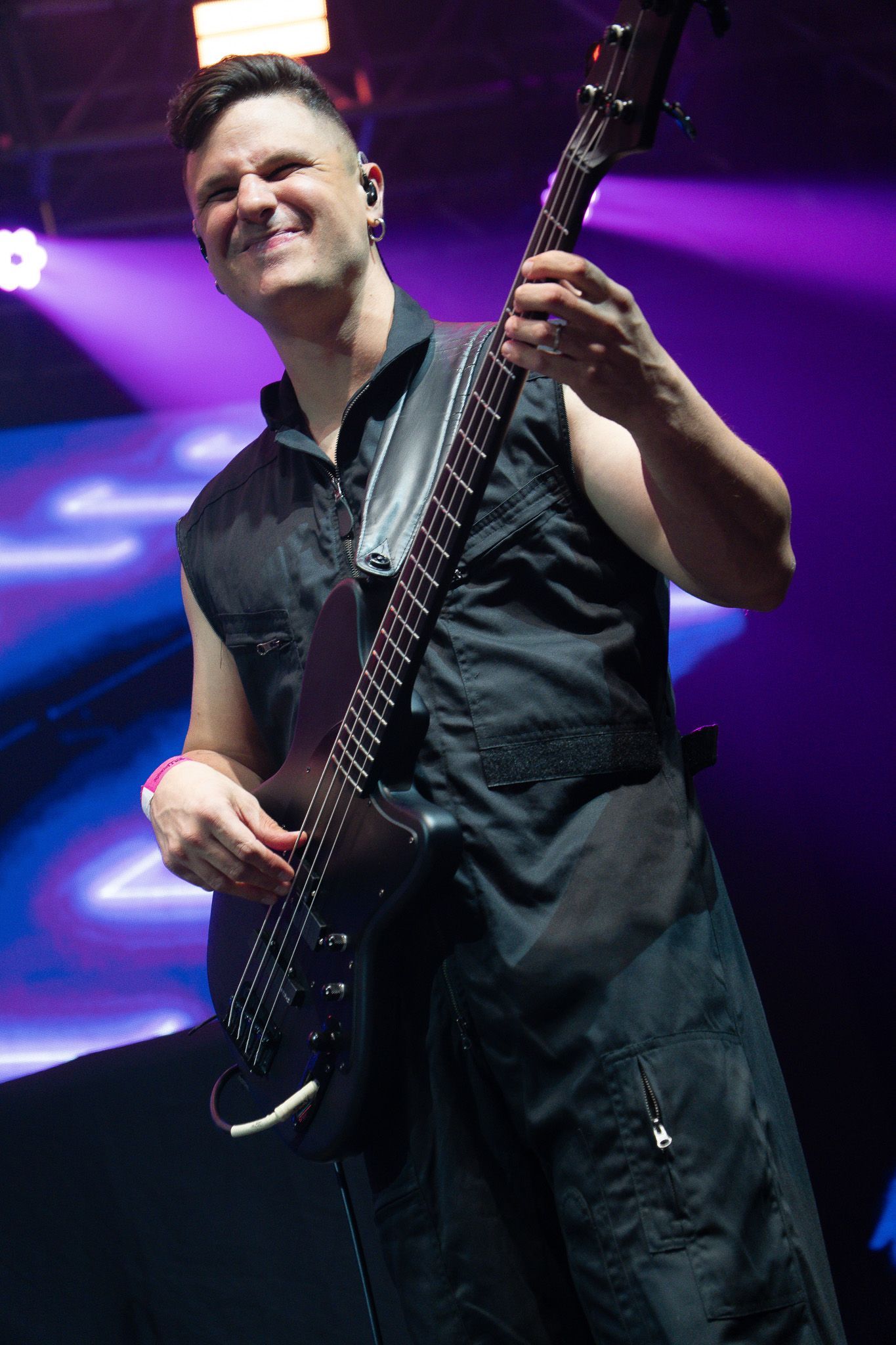 Um músico sorri enquanto se apresenta no palco com um baixo, iluminado por luzes de palco roxas.