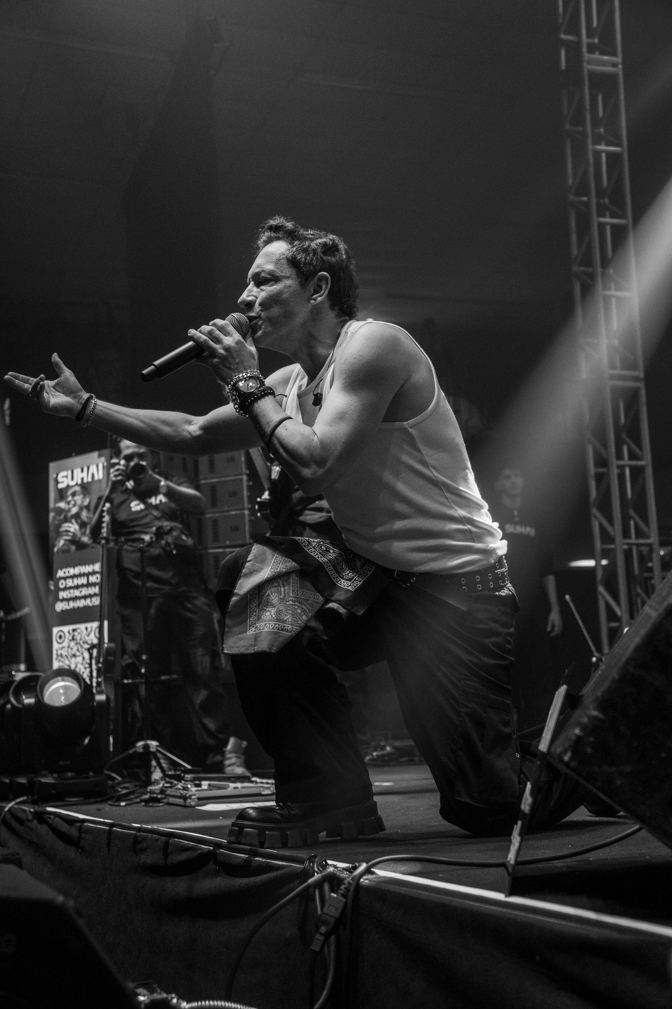 Um artista de regata ajoelha-se no palco, cantando ao microfone com um braço estendido sob um holofote brilhante.