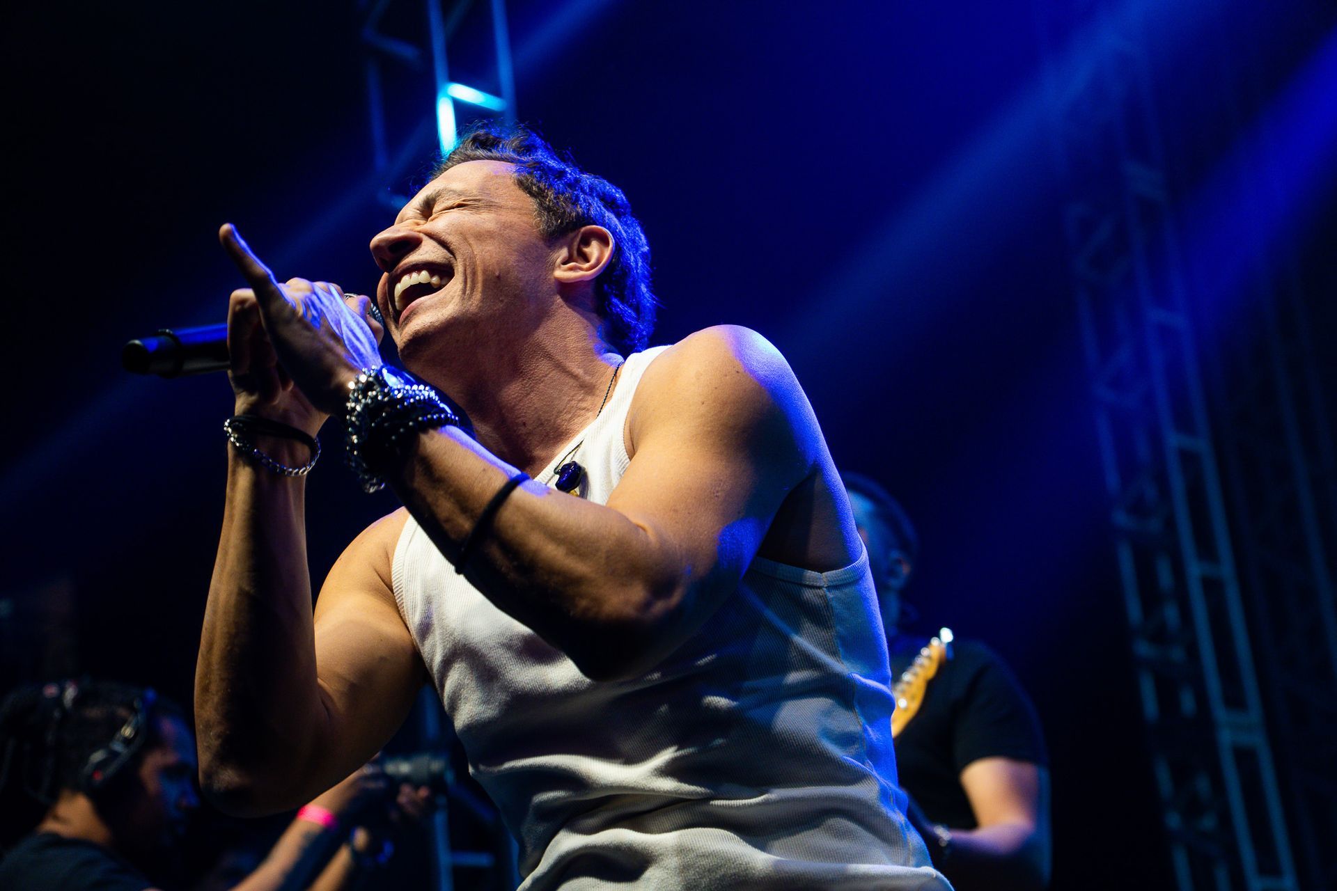 Um artista vestindo uma regata branca canta com entusiasmo em um microfone em um palco com iluminação azul de show.
