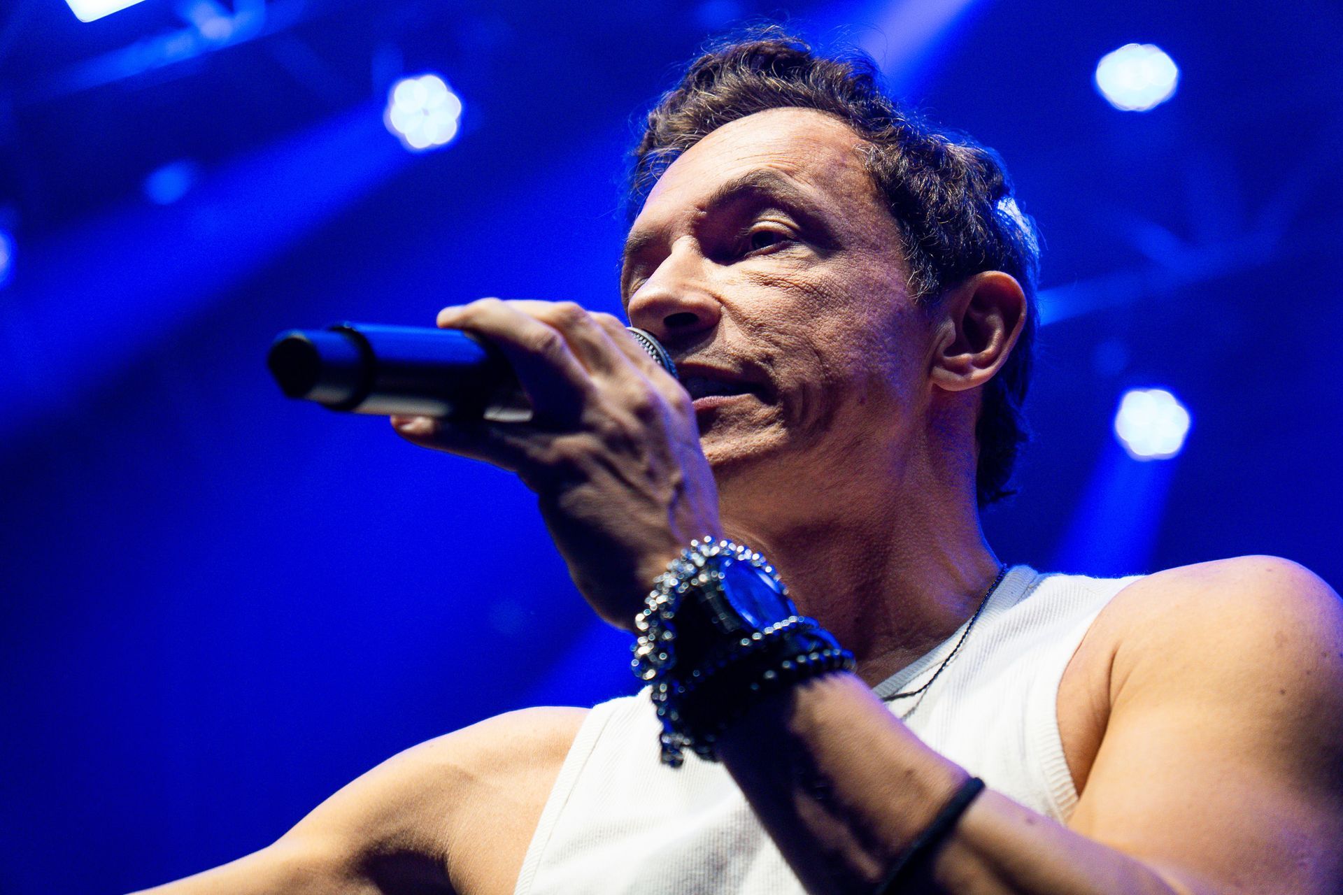 Um artista vestindo uma regata branca canta em um microfone sob luzes de palco azuis brilhantes.