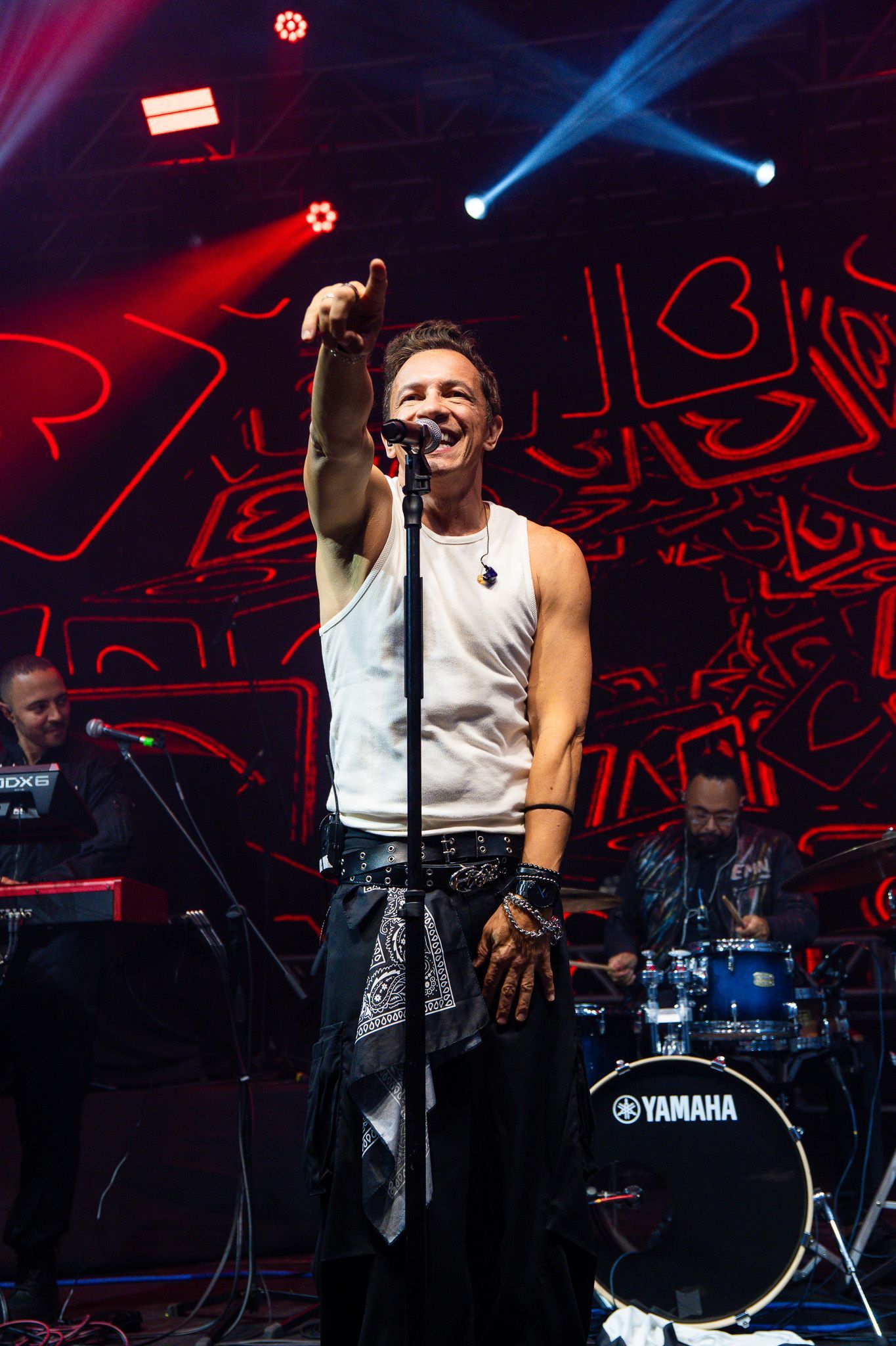 Um cantor se apresentando no palco com uma regata branca, apontando para cima, com um tecladista e um baterista ao fundo.