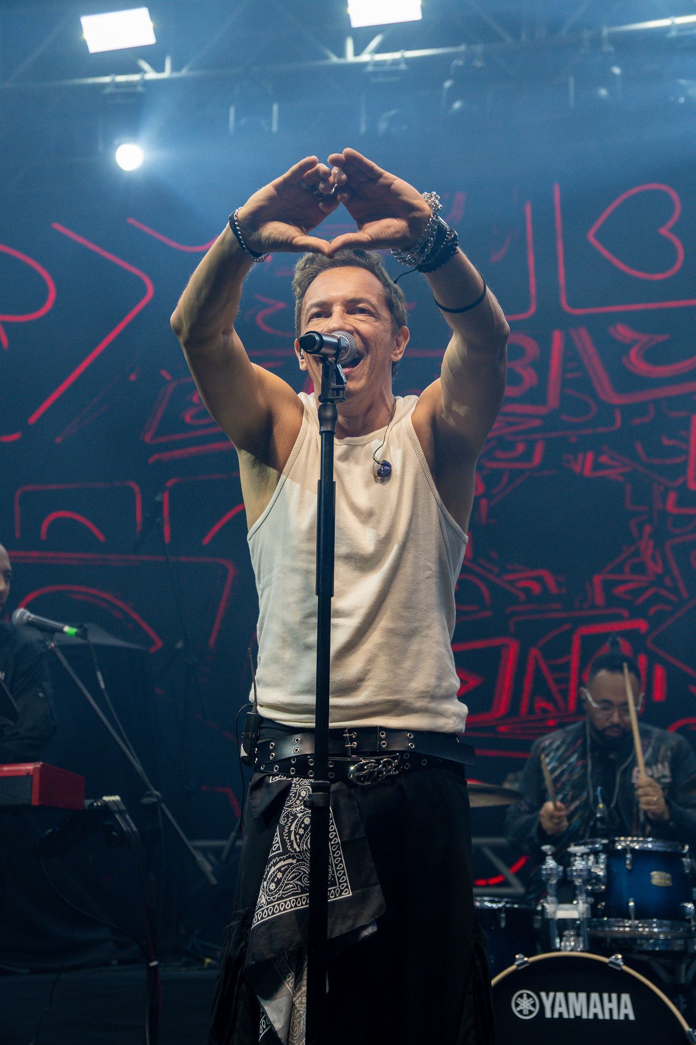 Um artista no palco, vestindo uma regata clara e calças escuras, forma um diamante com as mãos acima da cabeça.