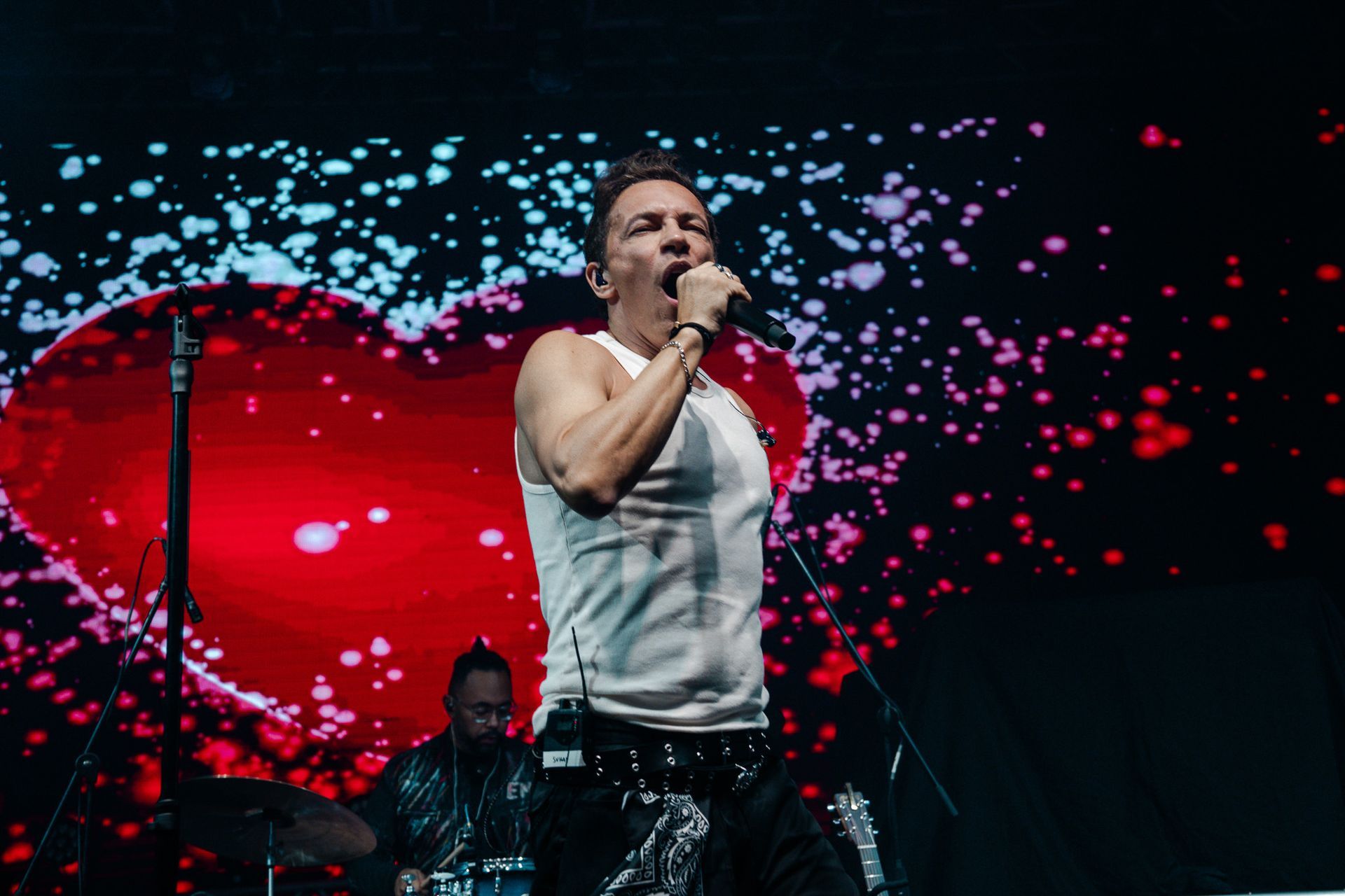 Um artista vestindo uma regata branca canta em um microfone no palco, tendo como fundo um grande coração vermelho brilhante.