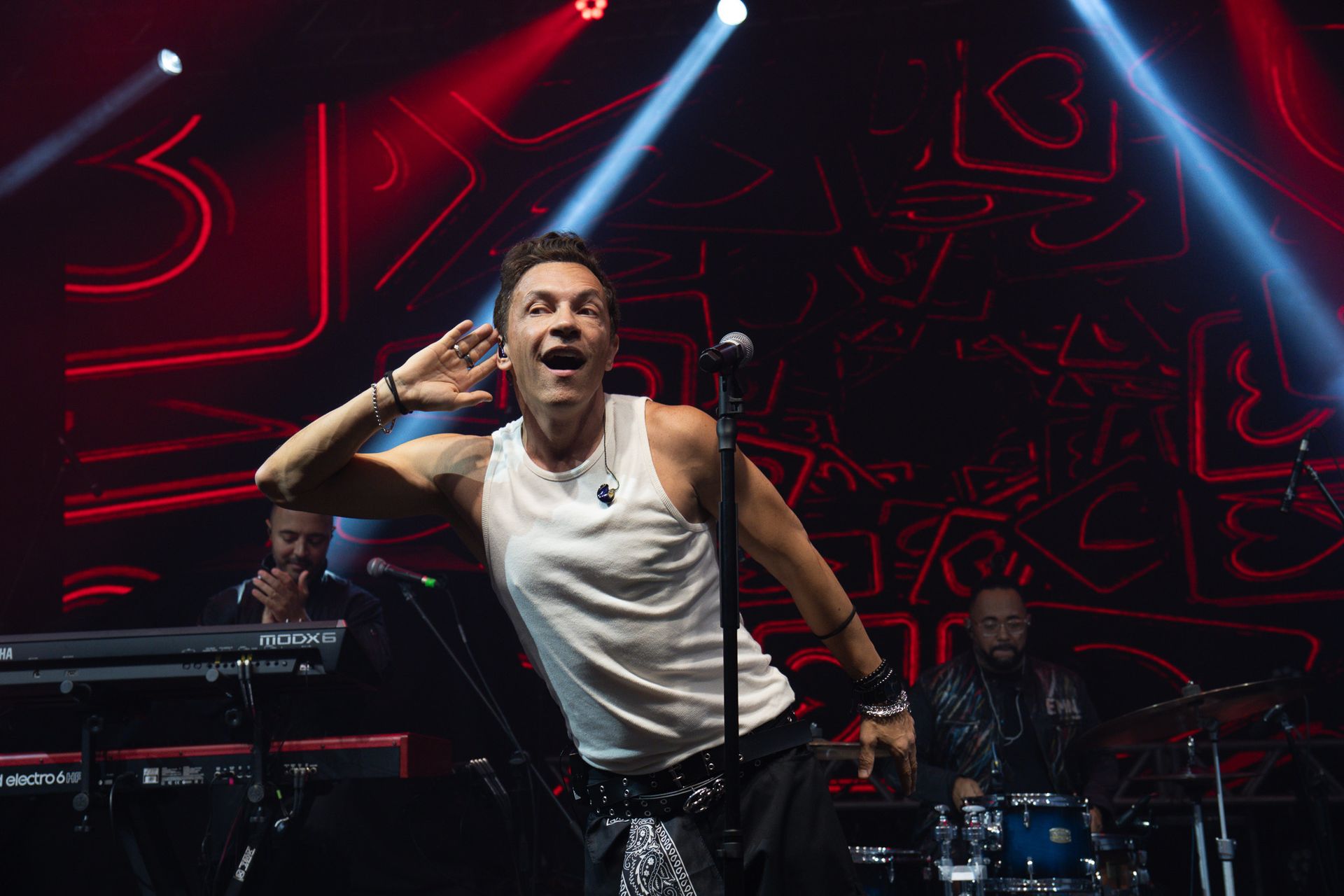 Um vocalista de regata branca inclina-se em direção à plateia com a mão na orelha, em um palco iluminado néon e com a banda.