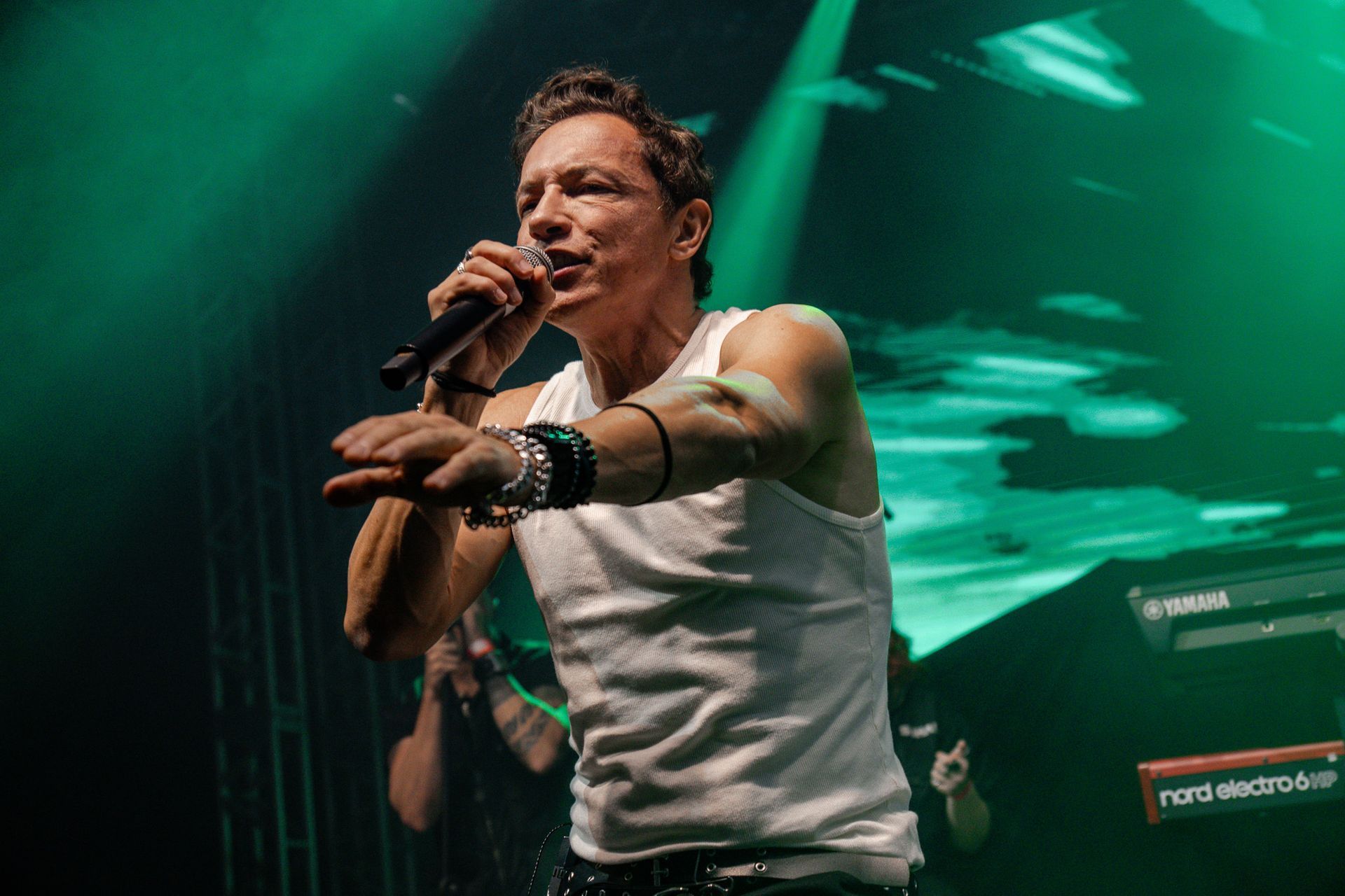 Um artista vestindo uma regata branca canta em um microfone no palco, gesticulando em direção à plateia.