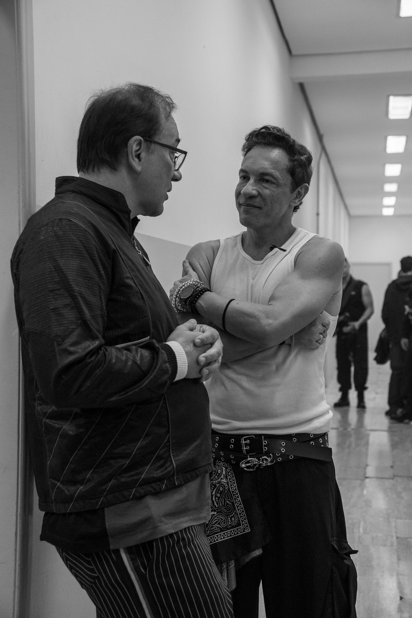 Duas pessoas em um corredor, uma vestindo uma jaqueta escura e a outra uma regata, conversando enquanto se encaram.