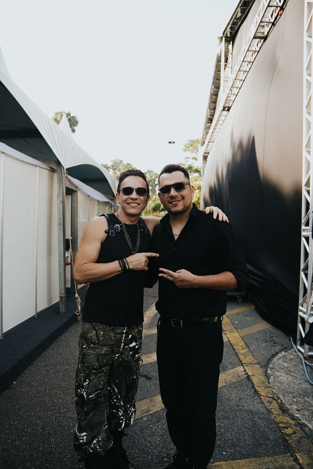 Dois homens posando ao ar livre. Um aponta, o outro estende a mão. Ambos usam óculos de sol e roupas pretas.