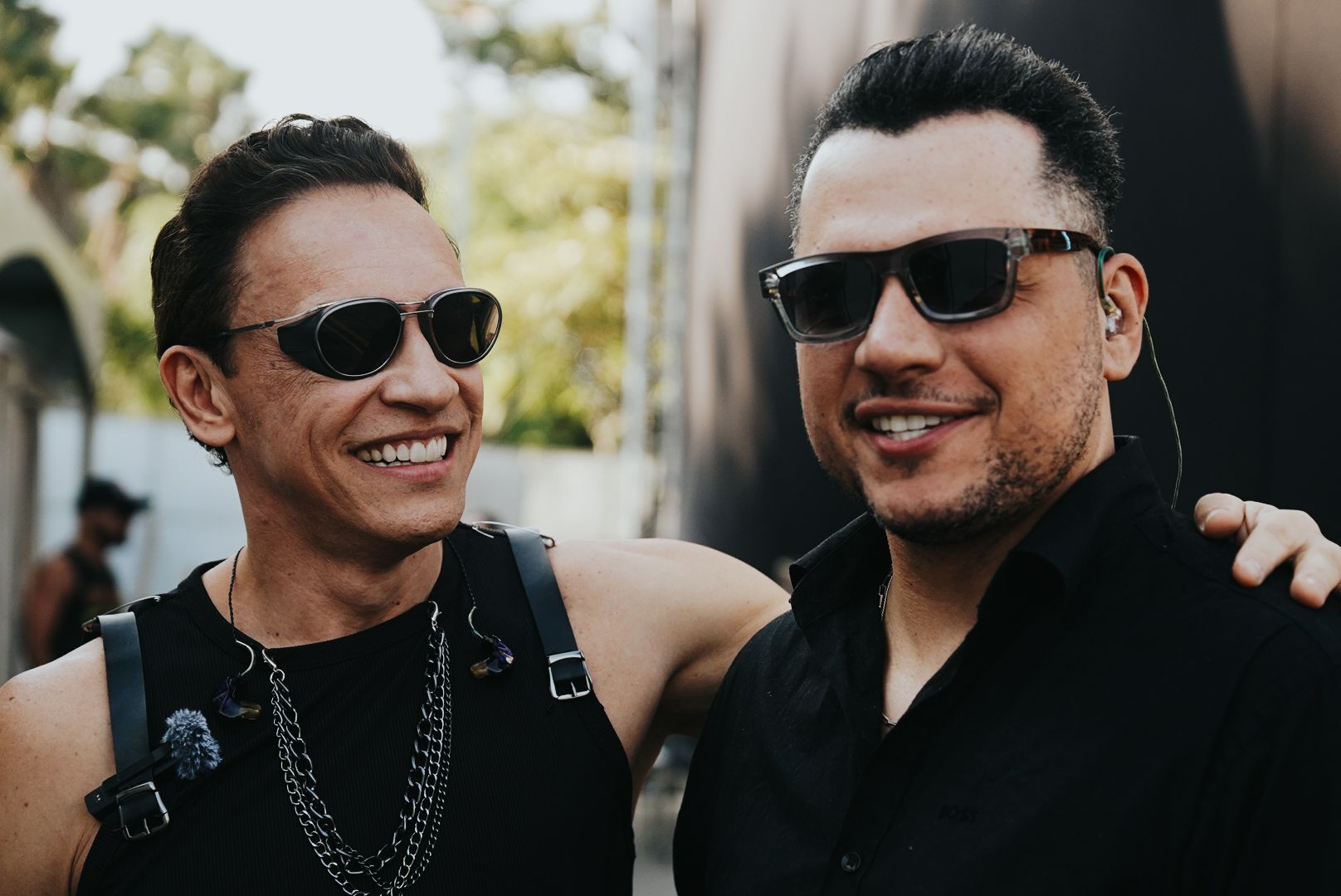 Dois homens sorridentes, usando óculos de sol, em pé ao ar livre. Um veste uma regata preta, o outro uma camisa preta.