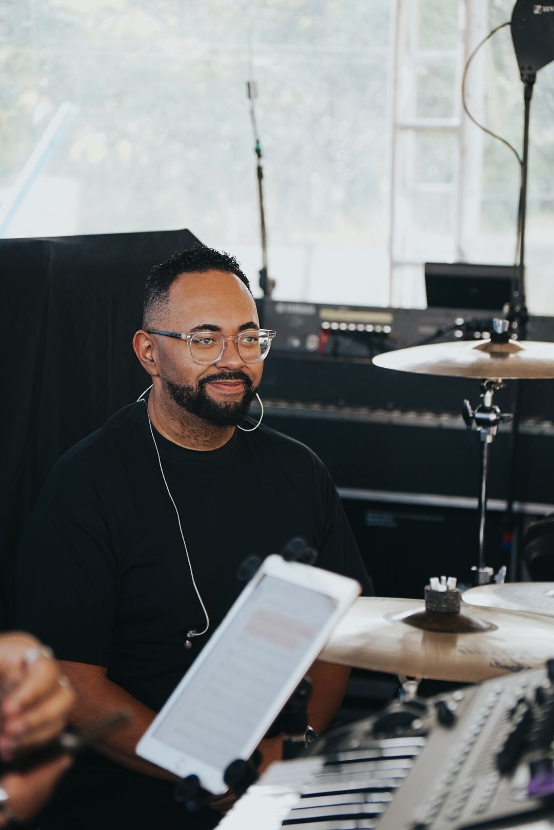 Um artista de óculos está sentado ao teclado, sorrindo. Um microfone e uma bateria estão visíveis.