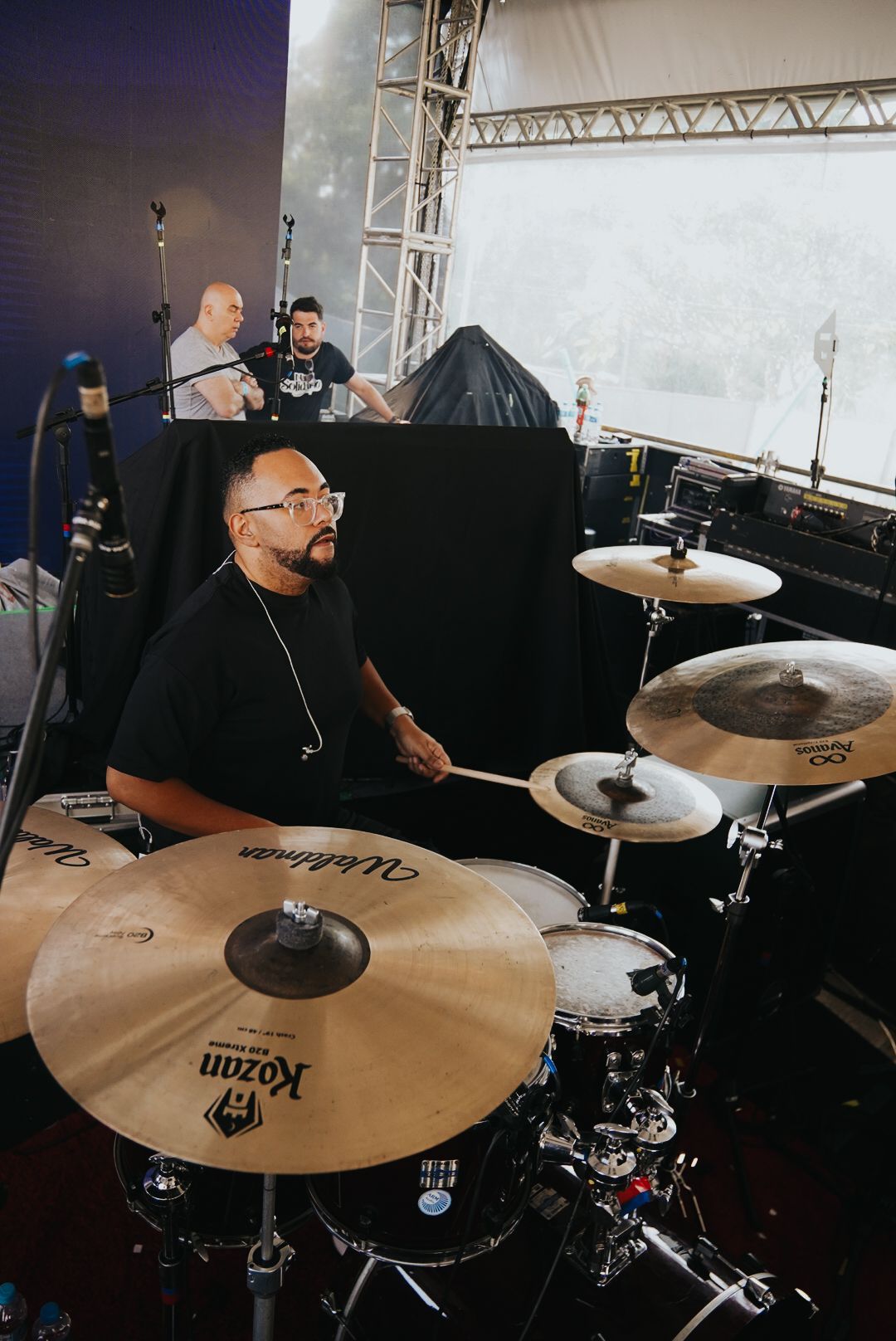 Baterista tocando bateria, com dois homens em pé atrás de uma superfície preta em um ambiente externo.