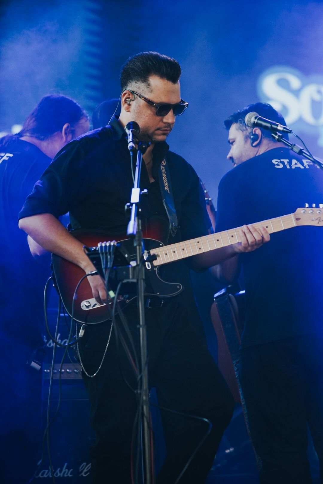 Guitarrista de camisa preta tocando uma guitarra vermelha e preta no palco, com dois membros da equipe atrás dele.