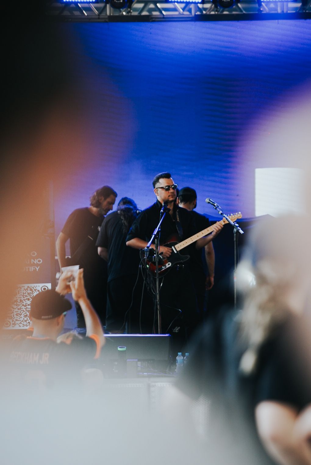 Banda se apresentando no palco com fundo azul, com foco no guitarrista.