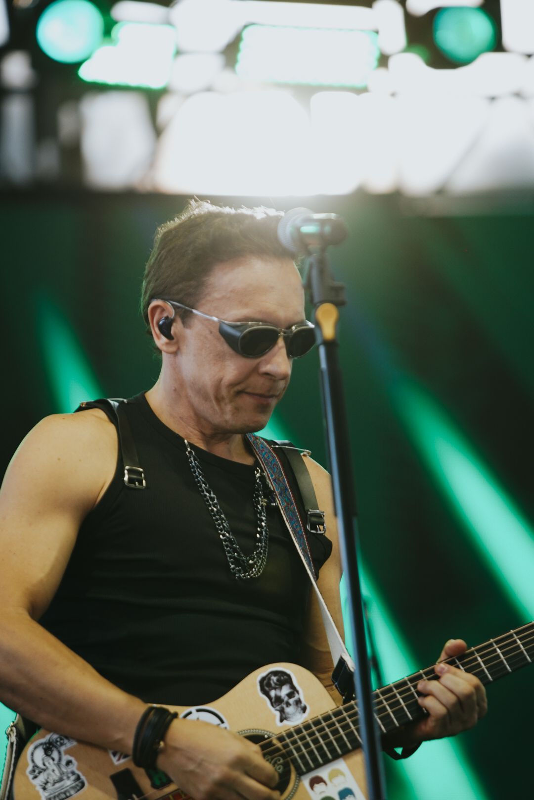 Um artista de óculos escuros toca violão no palco, vestindo uma regata preta e um colar.