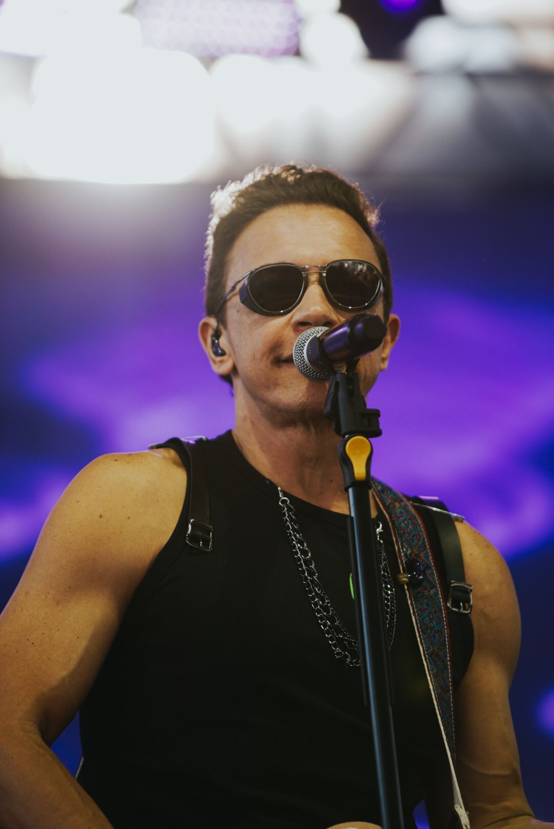 Um artista de óculos escuros cantando ao microfone, vestindo uma regata preta, no palco.
