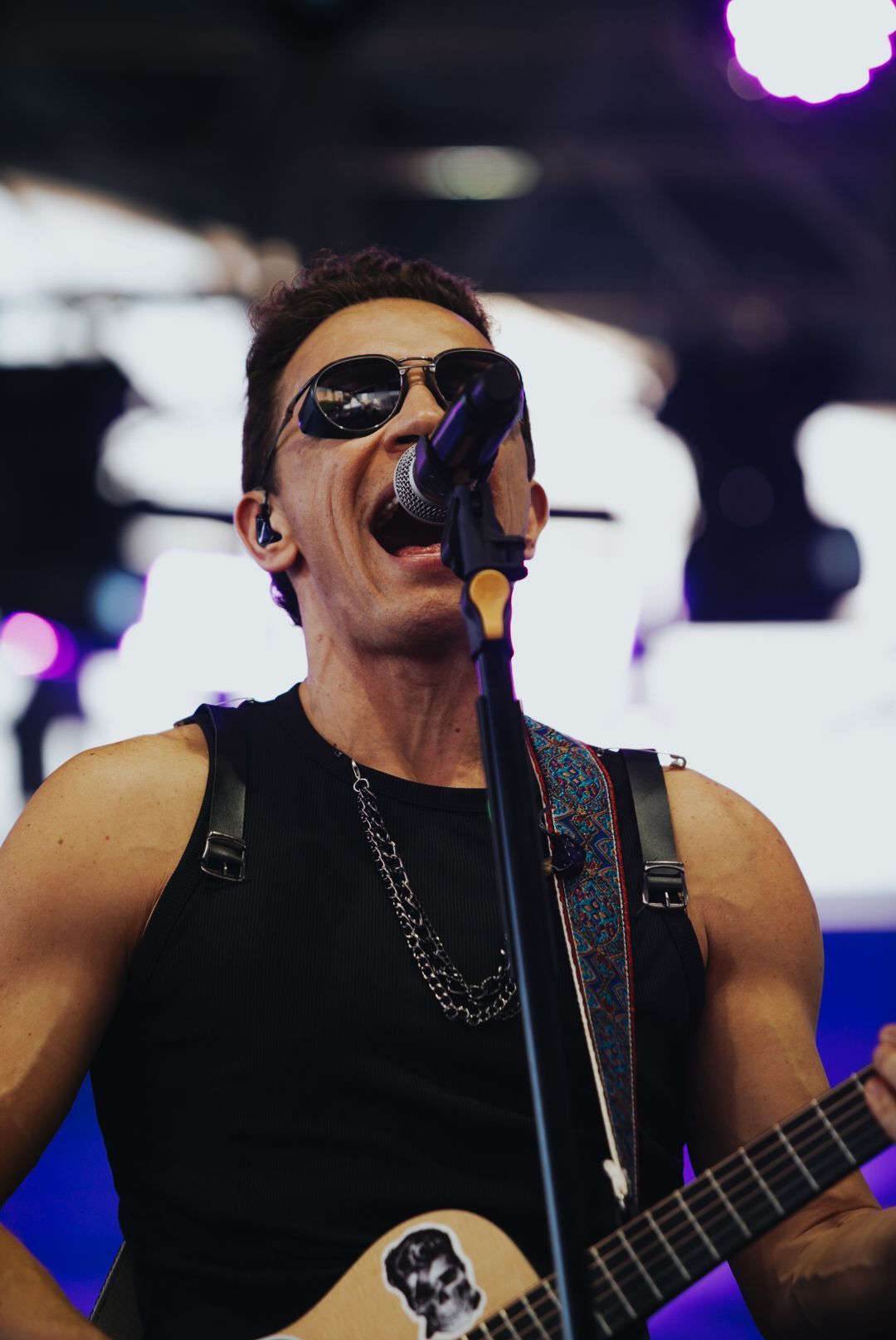 Um artista de óculos escuros cantando ao microfone enquanto toca violão no palco.