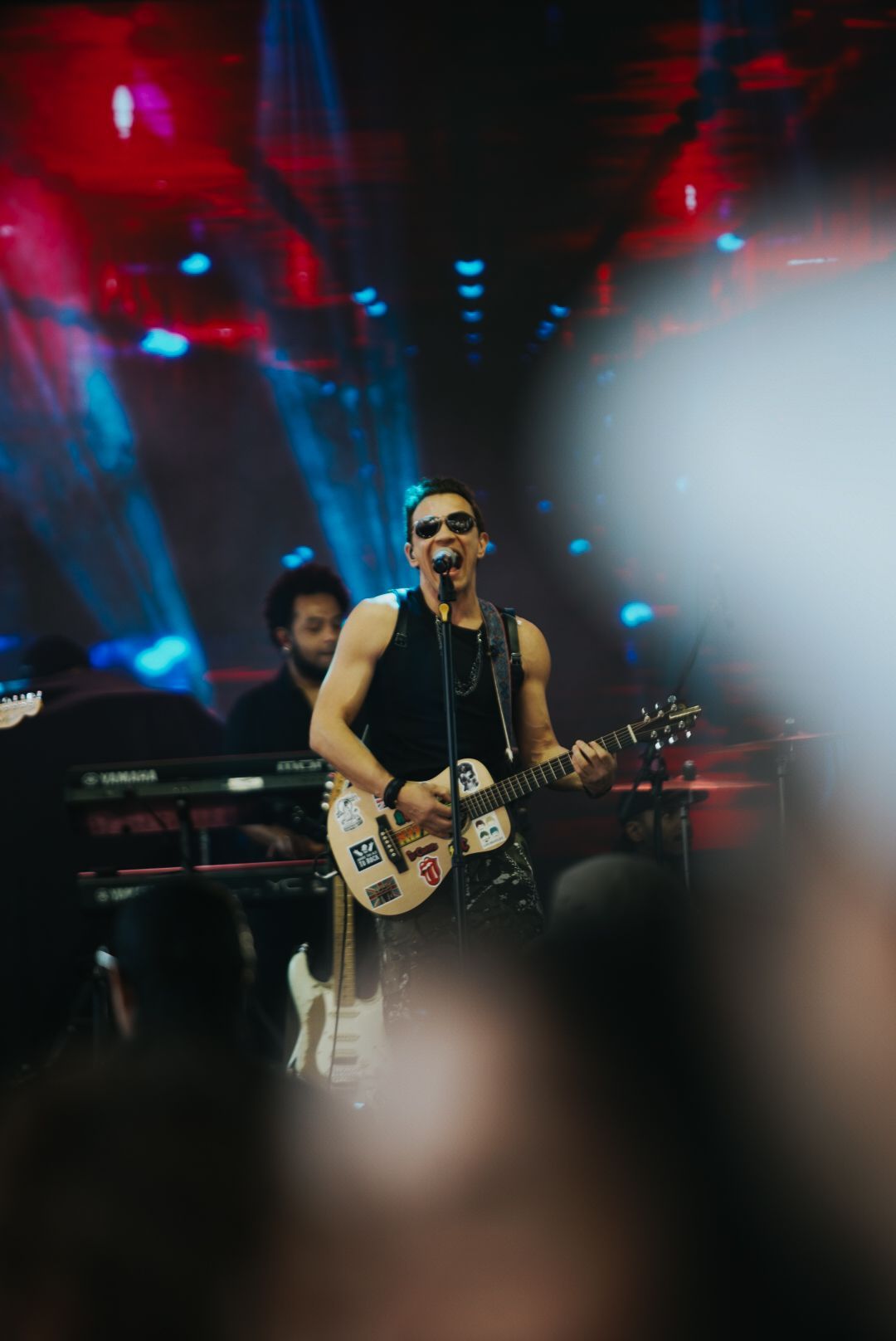 Um artista usando óculos escuros toca guitarra no palco, com luzes piscando em vermelho e azul.