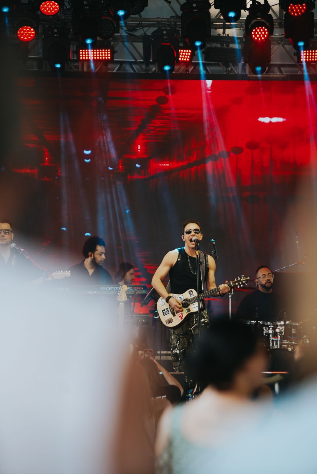 Banda se apresentando no palco com luzes vermelhas e azuis. O guitarrista canta e toca, com a plateia desfocada.