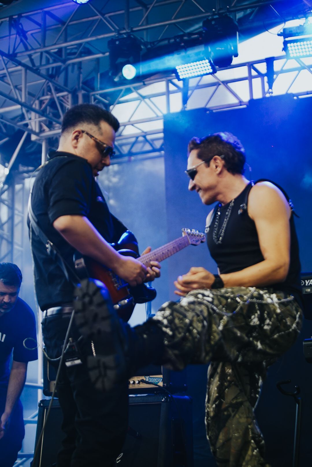 Dois músicos no palco, um tocando guitarra, o outro agachado, sorrindo. Iluminação azul.