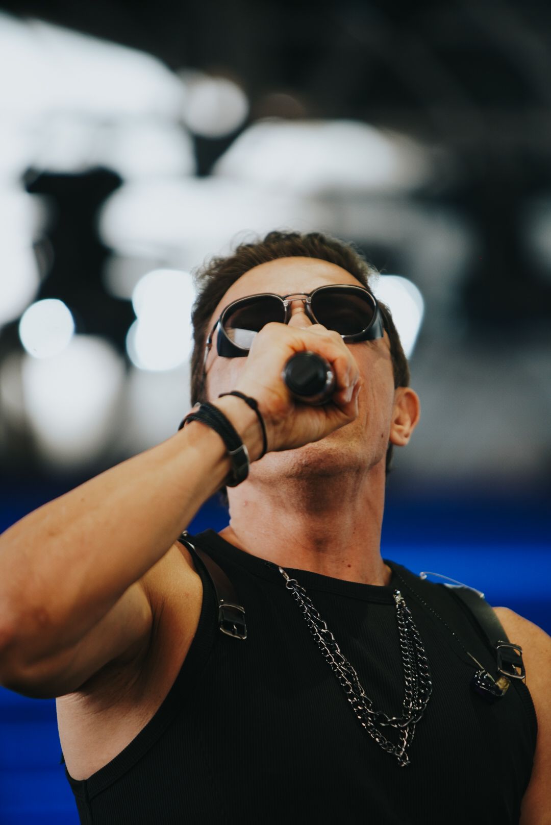 Um artista de óculos escuros e microfone canta, vestindo uma regata preta e um colar.