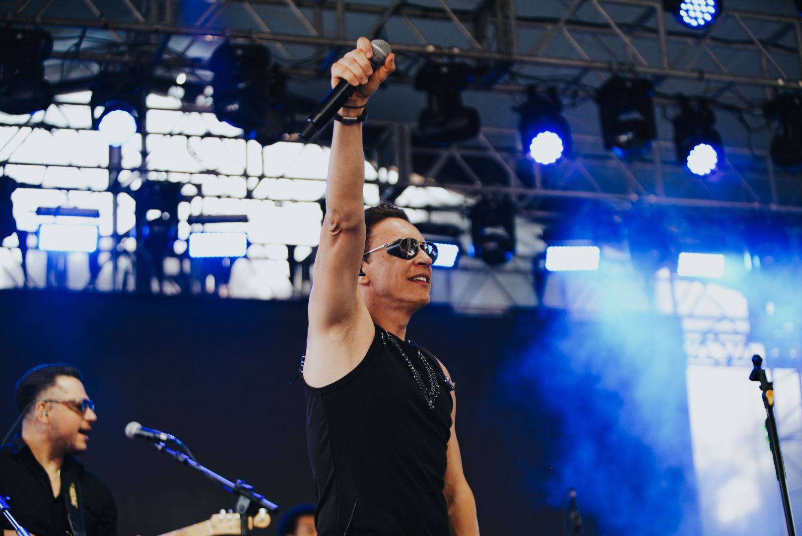 Cantor no palco segurando o microfone no alto; luzes azuis de palco ao fundo.