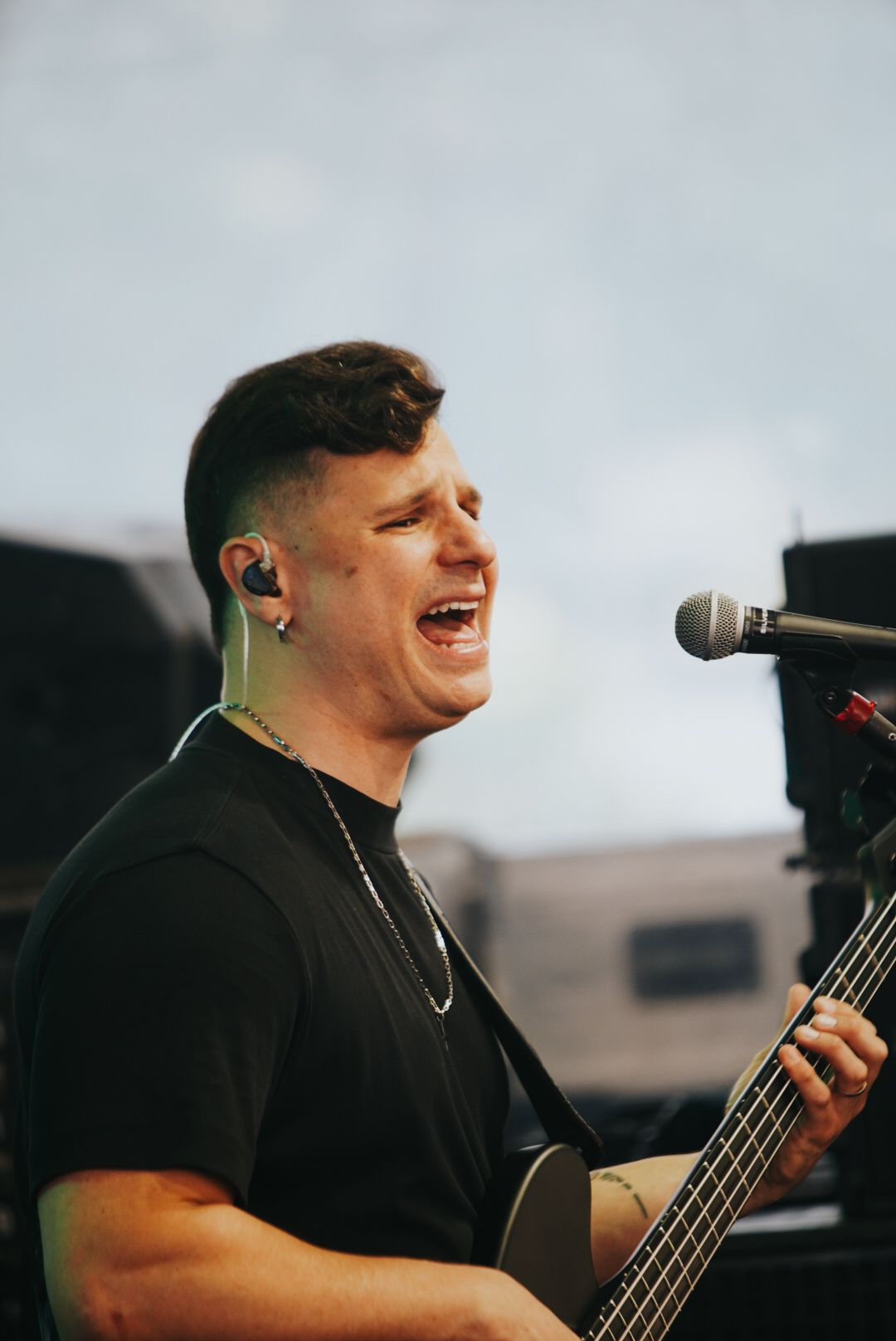 Um artista cantando e tocando baixo ao ar livre. Ele tem cabelo castanho curto e está vestindo uma camisa preta.