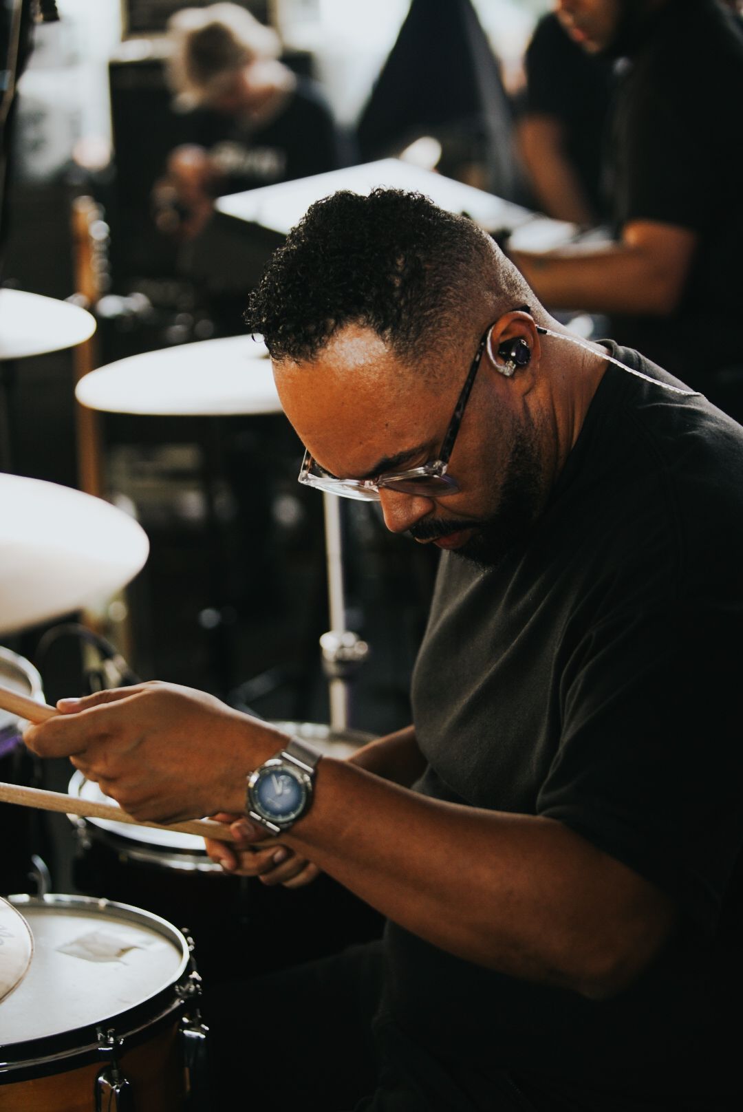 Baterista tocando bateria em um estúdio, usando óculos e relógio.