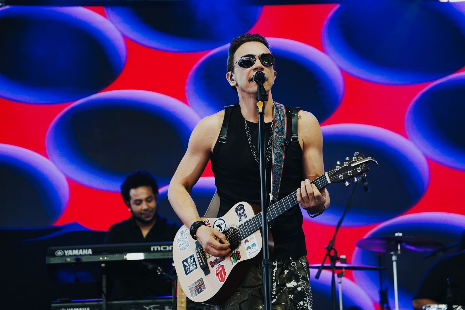 Um artista tocando guitarra no palco, usando óculos escuros, regata preta e calça camuflada, com um tecladista ao fundo.
