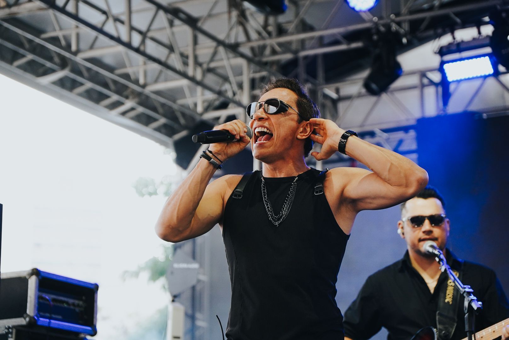 Um artista cantando em um microfone no palco, usando óculos escuros e uma regata preta. Um colega de banda aparece ao fundo.