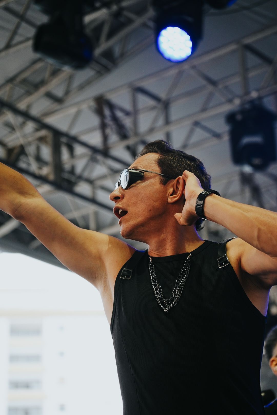 Um artista no palco, de óculos escuros e blusa preta sem mangas, com o braço levantado, cantando.