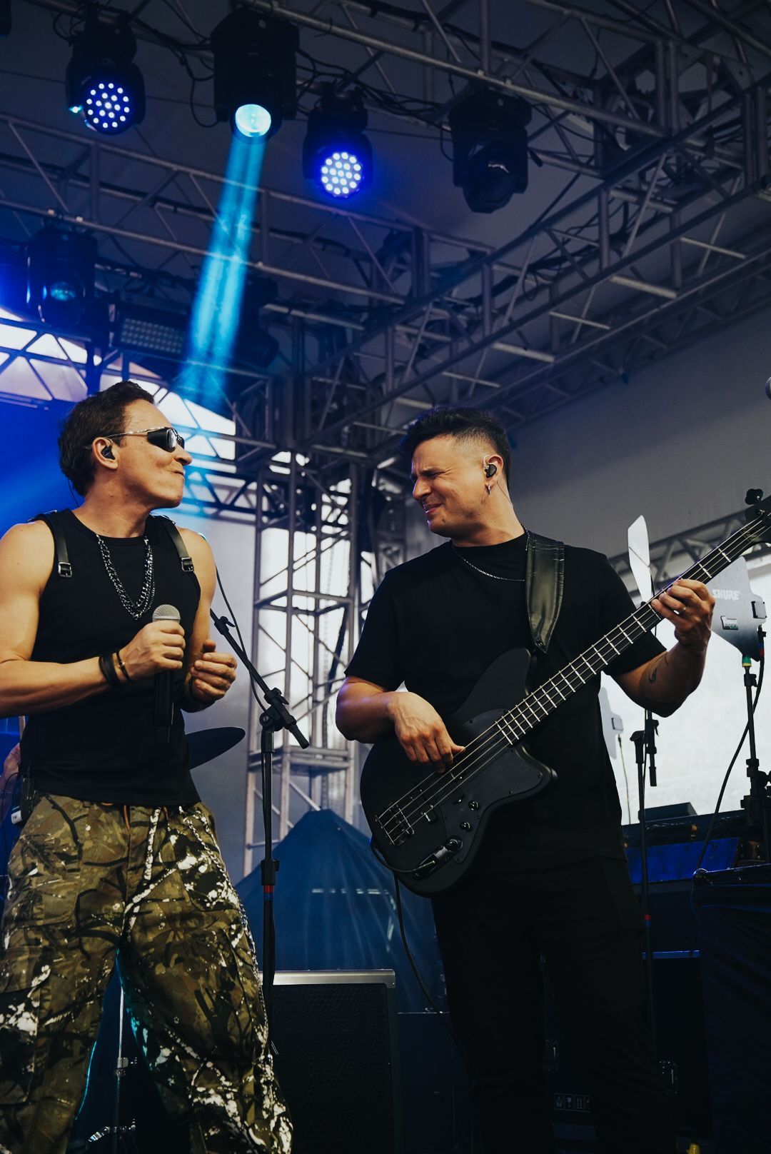 Dois artistas no palco: um cantando ao microfone, o outro tocando baixo. Luzes de palco brilhantes iluminam os artistas.