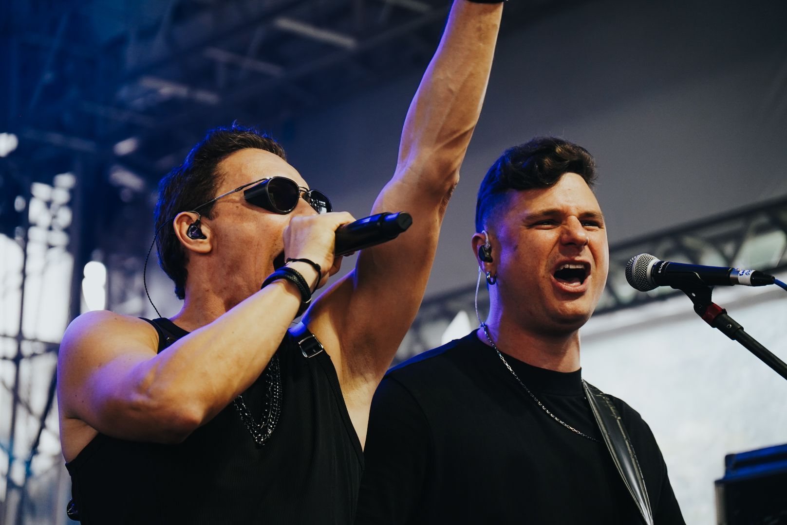 Dois artistas cantando no palco durante um show. Um deles levanta o braço e usa óculos escuros. O outro canta ao microfone.