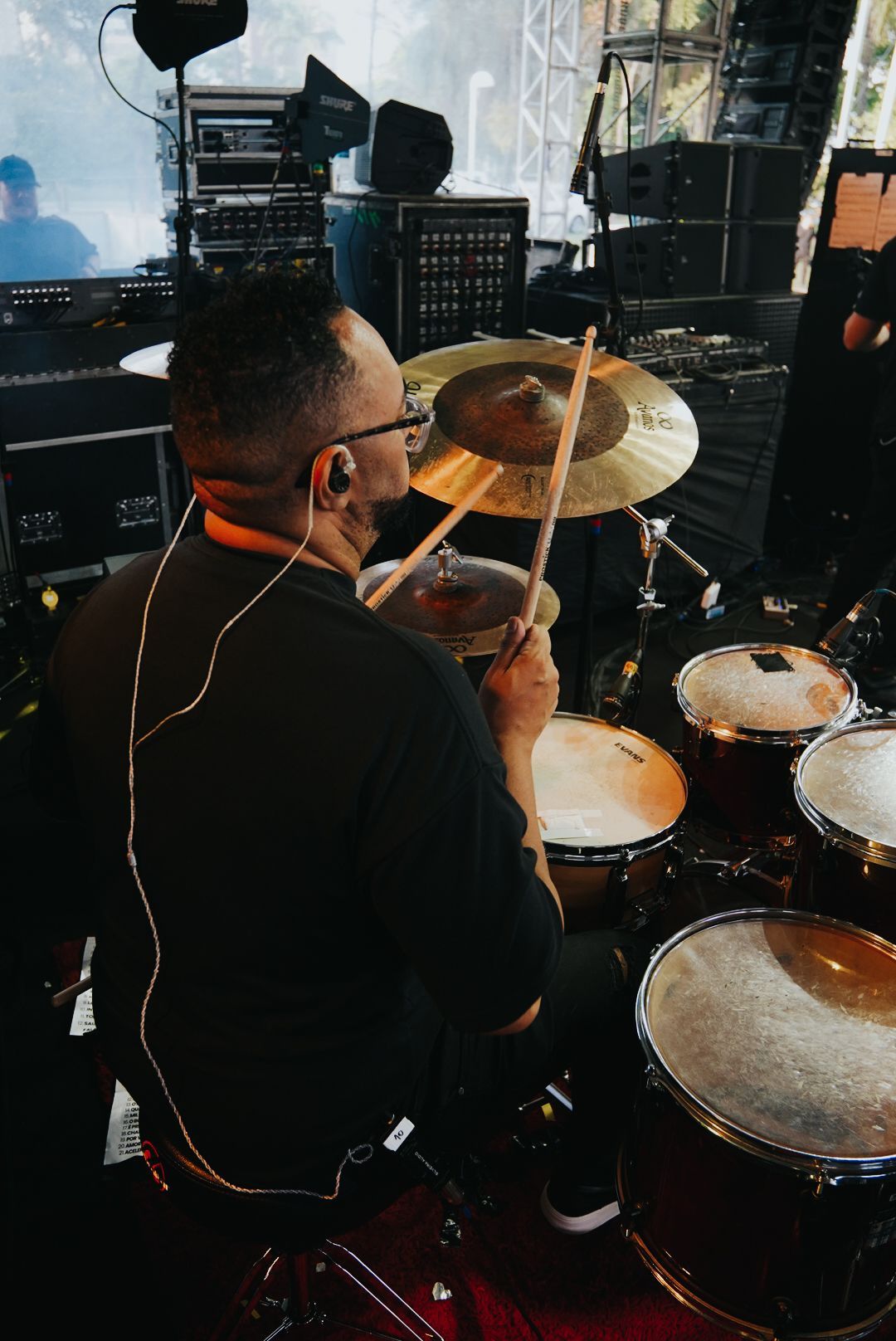 Baterista tocando bateria no palco. Ele está vestindo uma camisa preta e óculos.