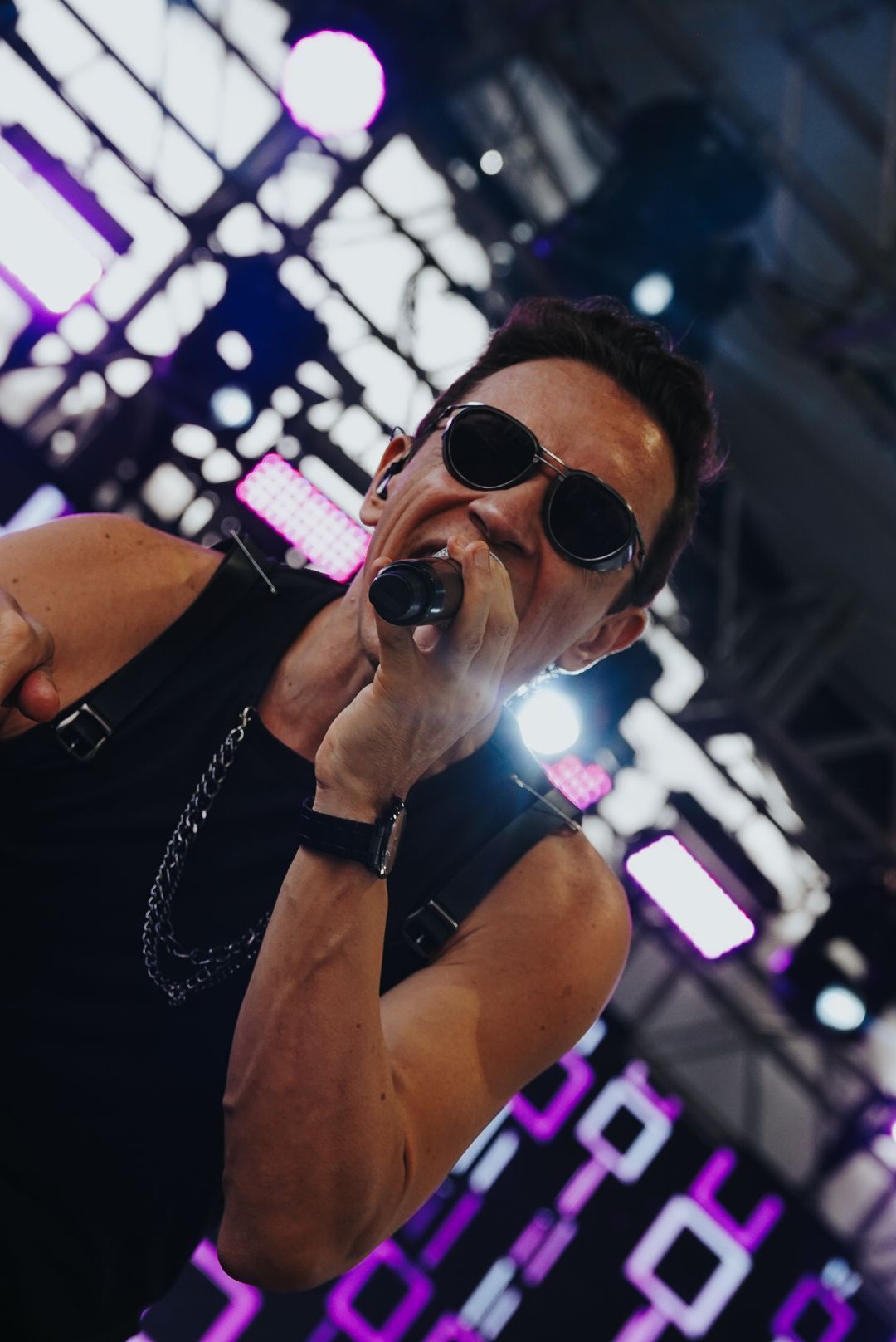 Cantor se apresentando no palco, usando óculos escuros e roupa preta, cantando em um microfone.