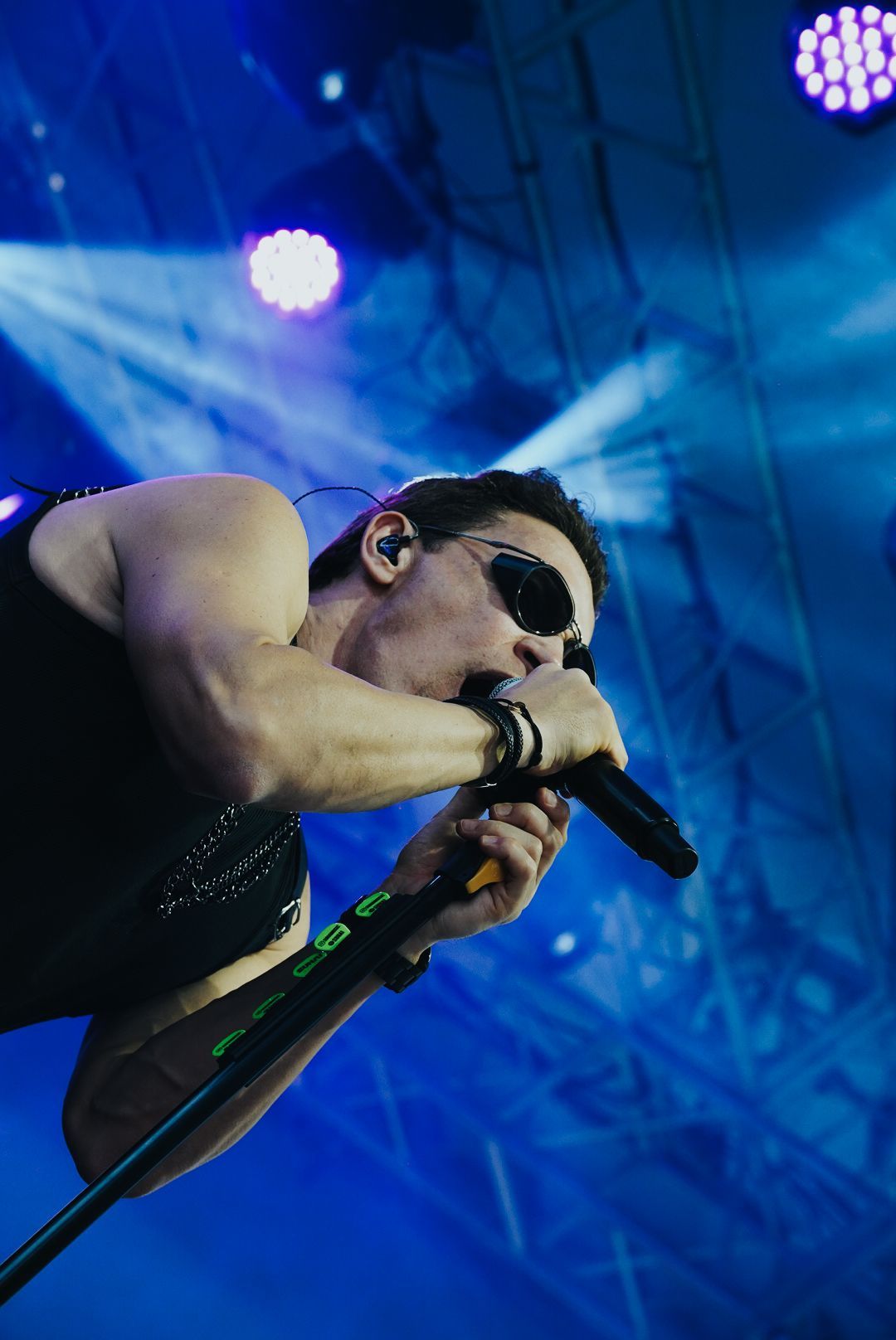 Um artista cantando ao microfone no palco, usando óculos escuros, regata preta e pulseiras. Iluminação de palco azul brilhante.