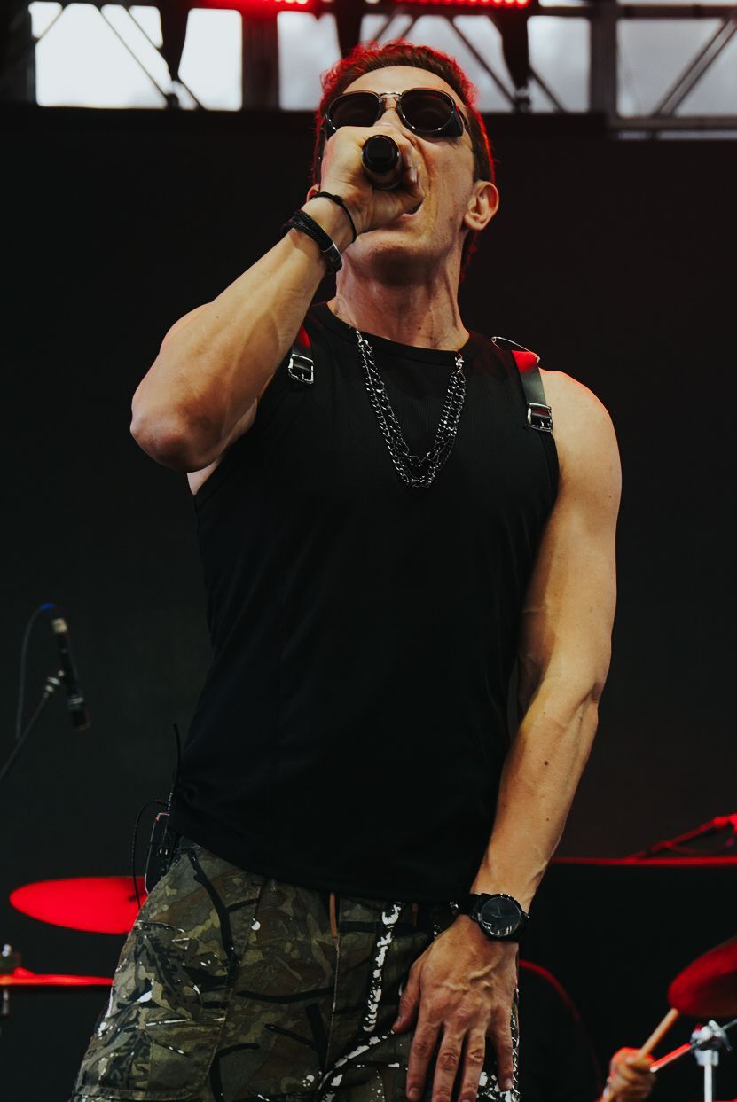 Um artista cantando em um microfone no palco, usando óculos escuros, regata preta e calça camuflada.