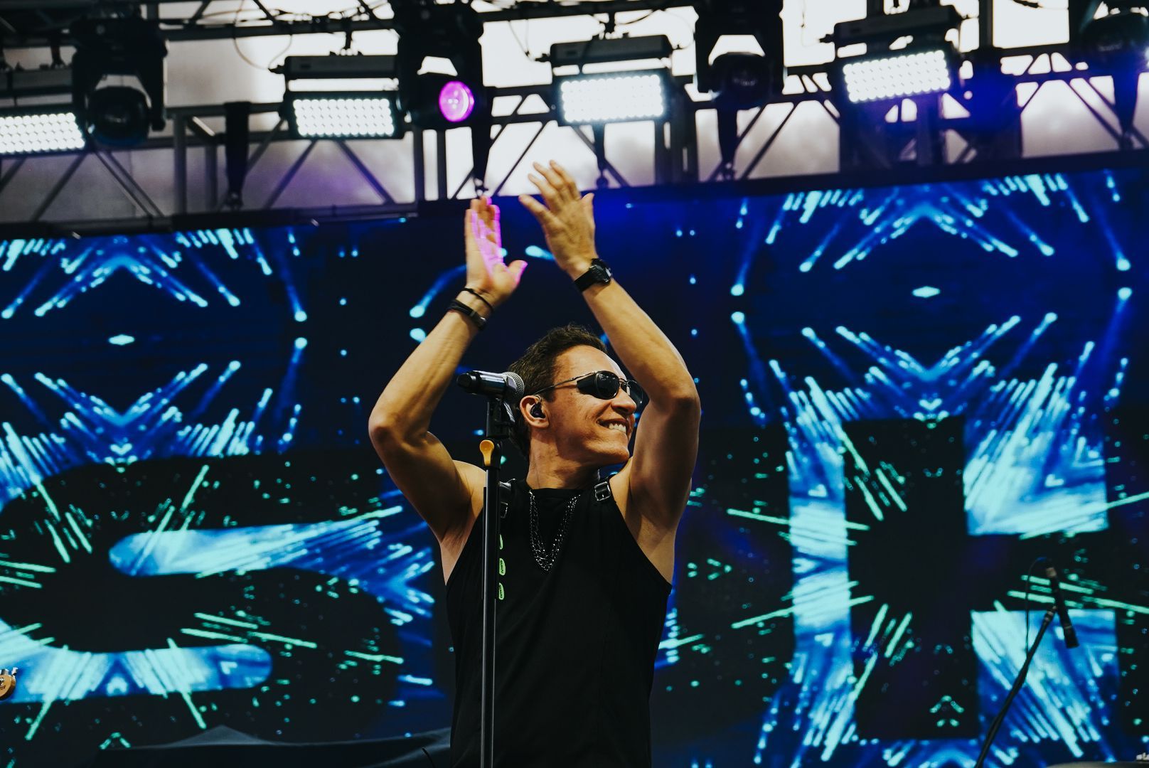 Um artista de óculos escuros aplaudindo no palco com luzes azuis ao fundo.