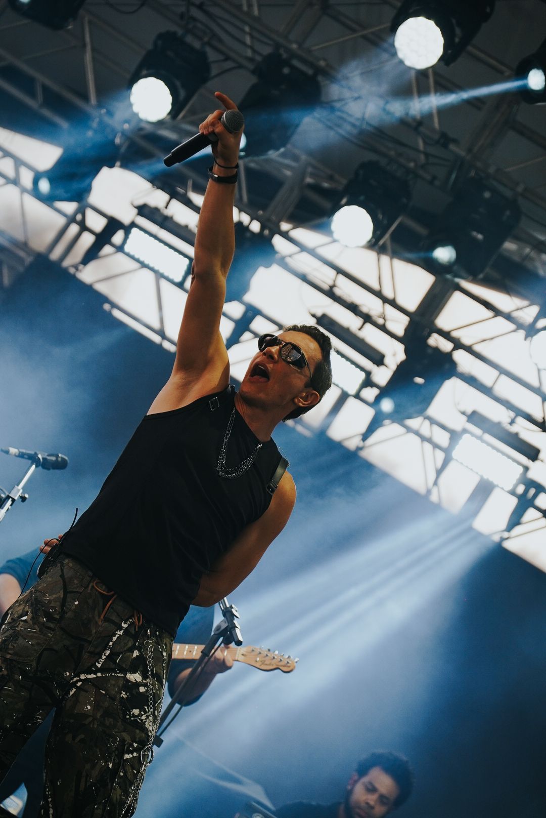 Cantor no palco com microfone, braço levantado, usando óculos escuros e colete preto, sob holofotes e com a plateia ao fundo.
