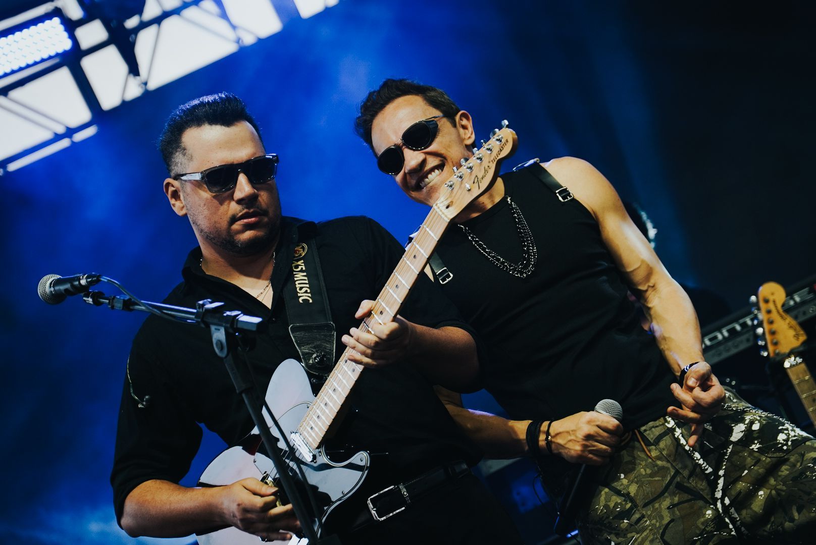 Dois artistas no palco: um toca guitarra elétrica, o outro canta ao microfone. Ambos usam óculos escuros.