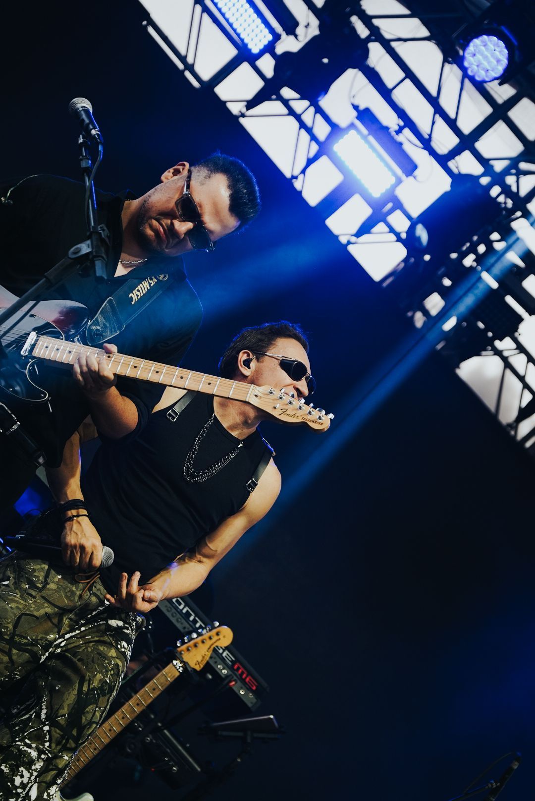 Dois músicos no palco, um tocando guitarra e o outro segurando um microfone. Luzes de palco azuis.