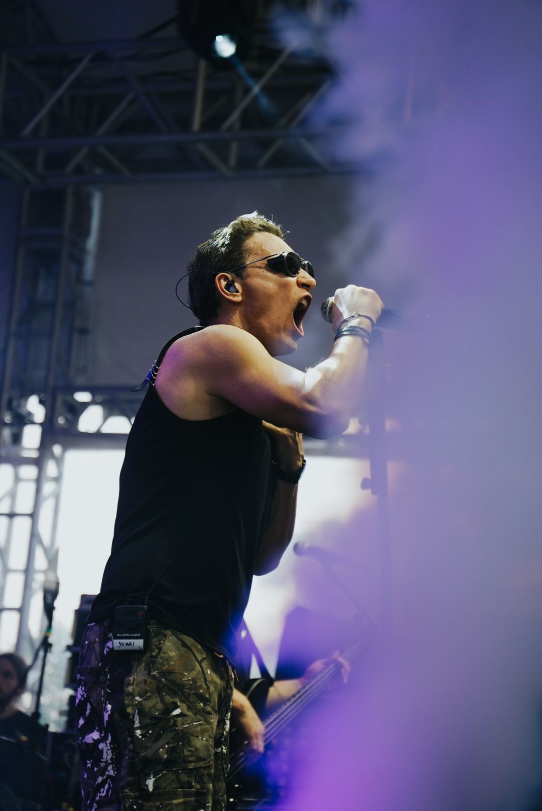 Um artista cantando de boca aberta, usando óculos escuros, regata preta e calça camuflada; iluminação de palco e fumaça.