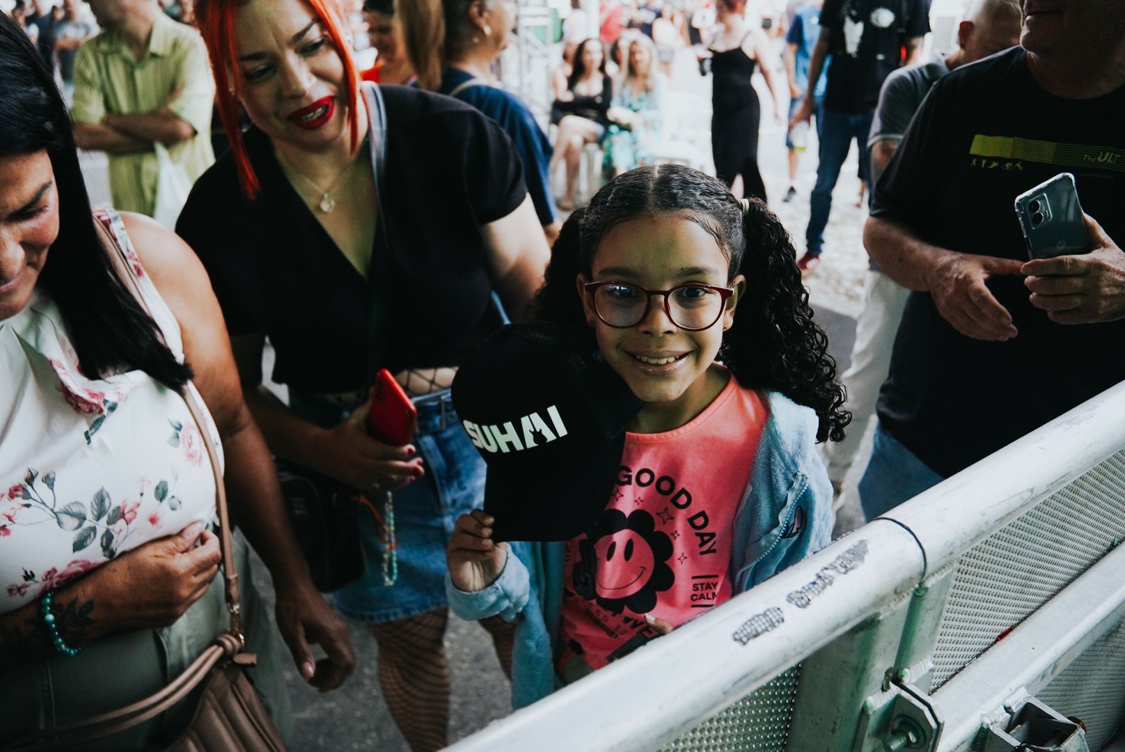 Menina de óculos sorri, segurando um chapéu, perto de uma barreira de metal, cercada por pessoas ao ar livre.
