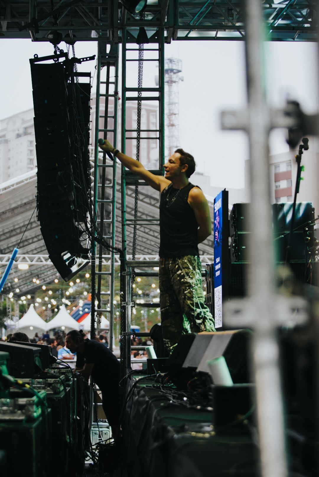 Um artista gesticulando em direção a um conjunto de alto-falantes, em pé no palco durante um evento ao ar livre.