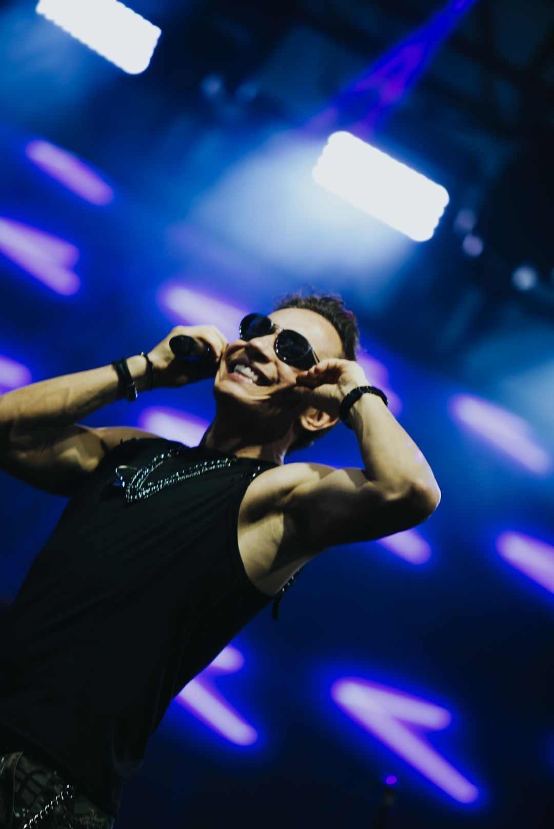 Um artista usando óculos escuros e blusa preta sem mangas, sorrindo e segurando as orelhas, em um palco com luzes azuis.