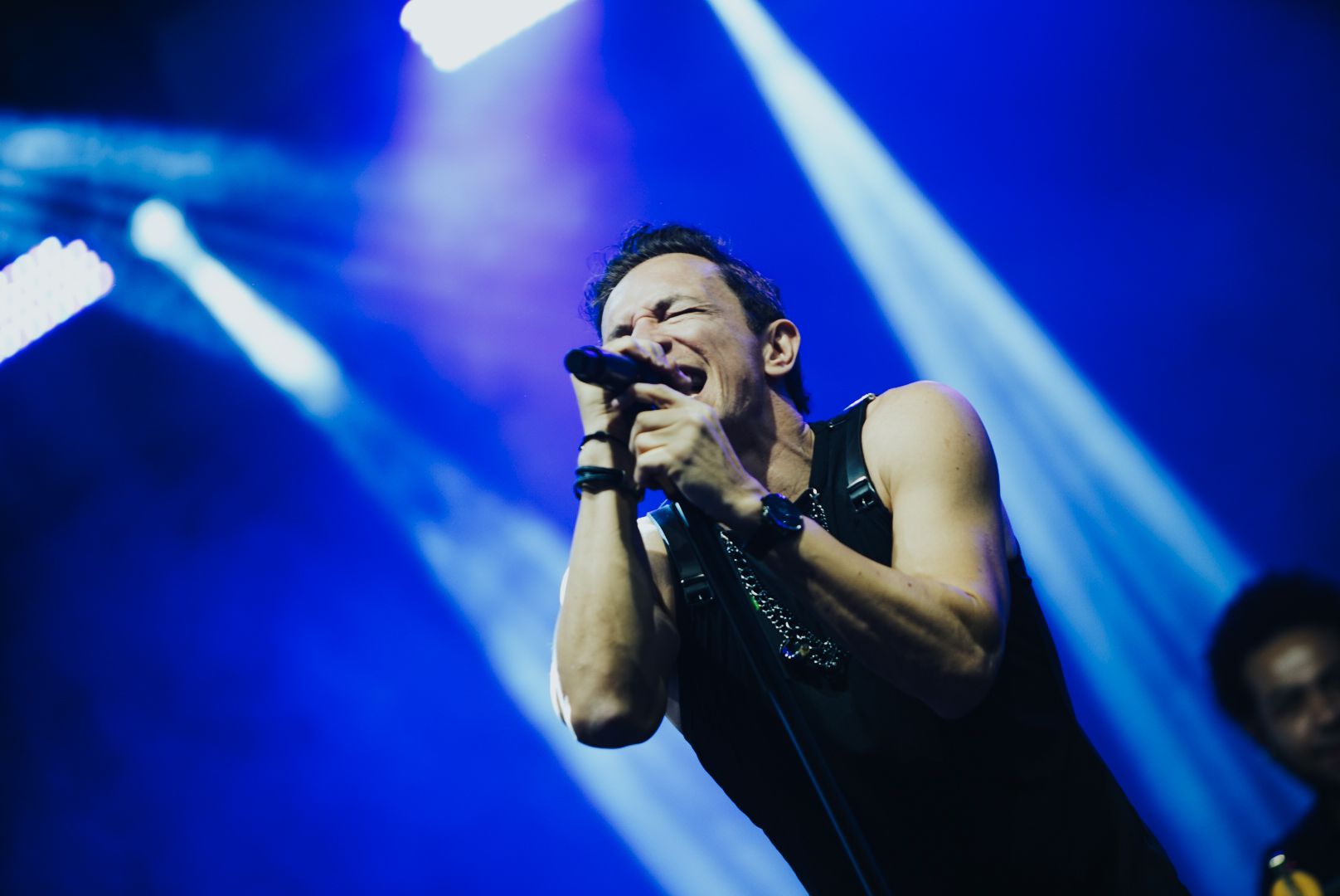 Um cantor se apresenta no palco, banhado por luz azul. Ele segura um microfone e canta de boca aberta.